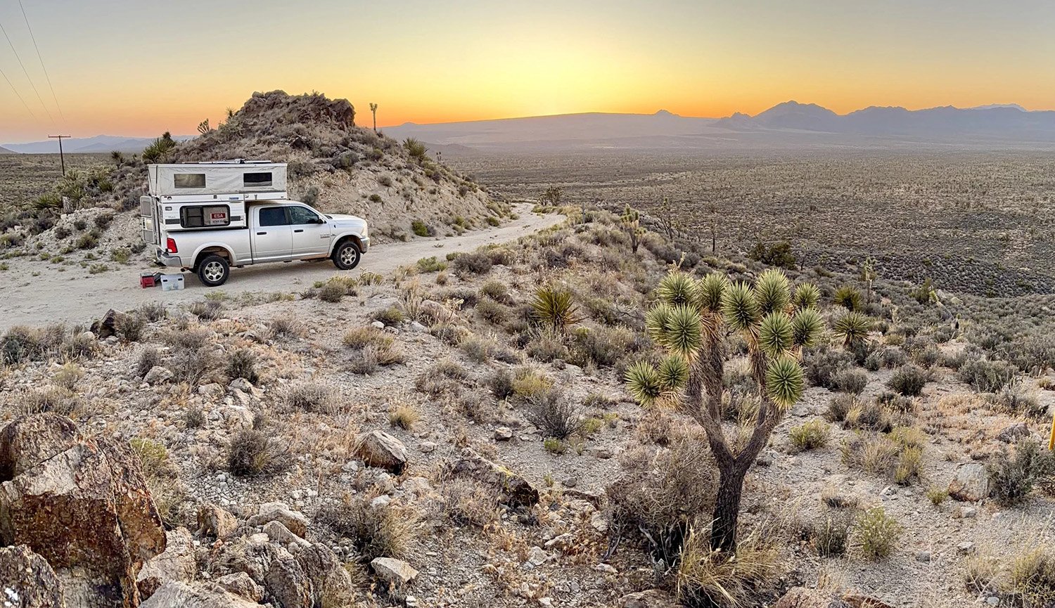 A white pickup truck with a camper shell parked on a dirt roadside in a desert landscape at sunset. The scene includes sparse desert vegetation, a Joshua tree, rocky terrain, and distant mountains under a colorful sky.