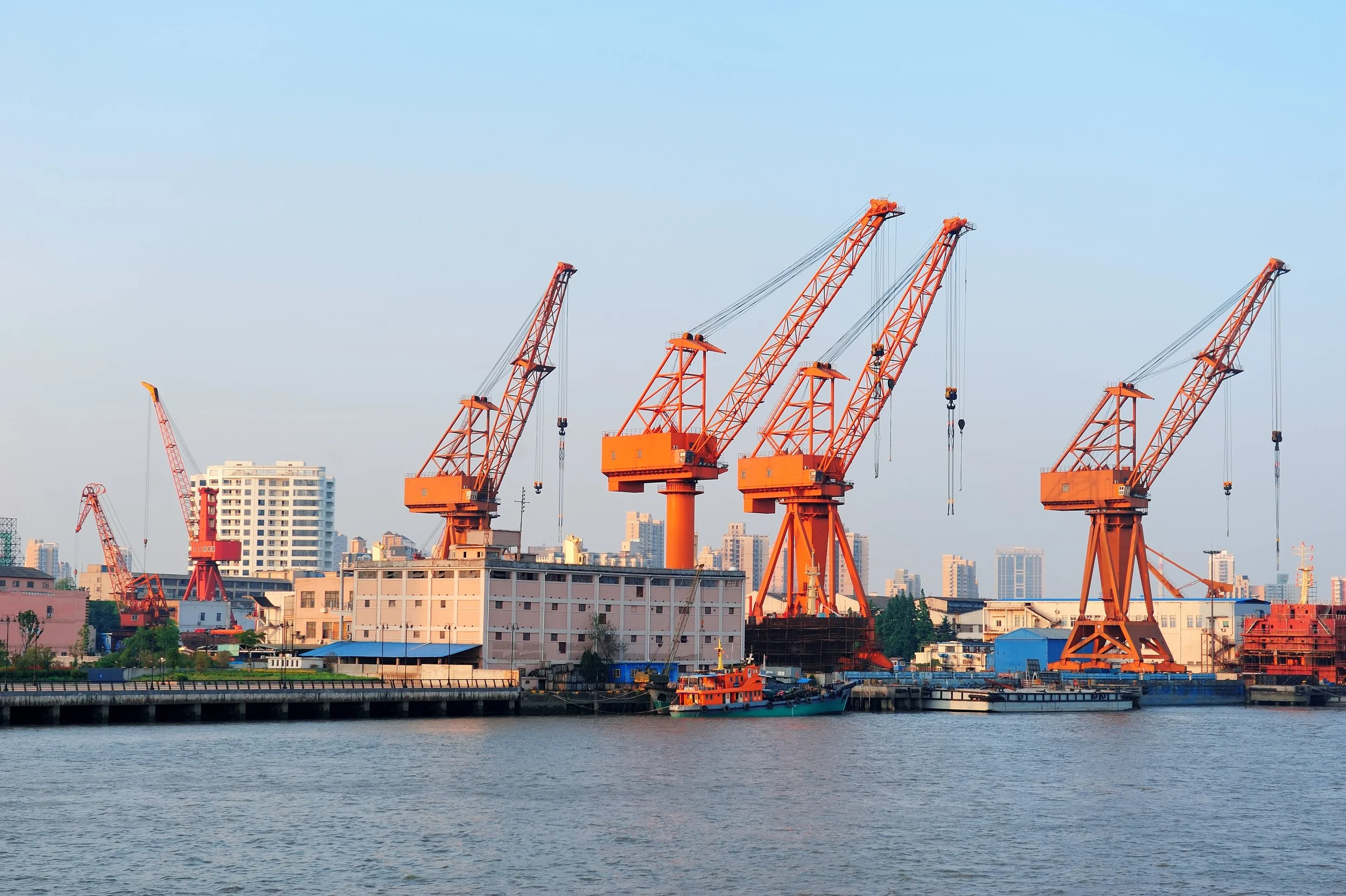 View of an industrial harbor with multiple large orange cranes and a boat in the water, with city buildings in the background under a clear blue sky.