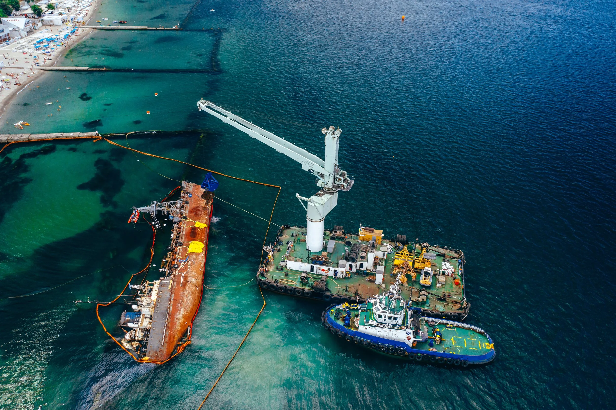 Aerial view of a large ship being repaired in a port, with various boats and water surrounding it, and a beach with umbrellas and people nearby.