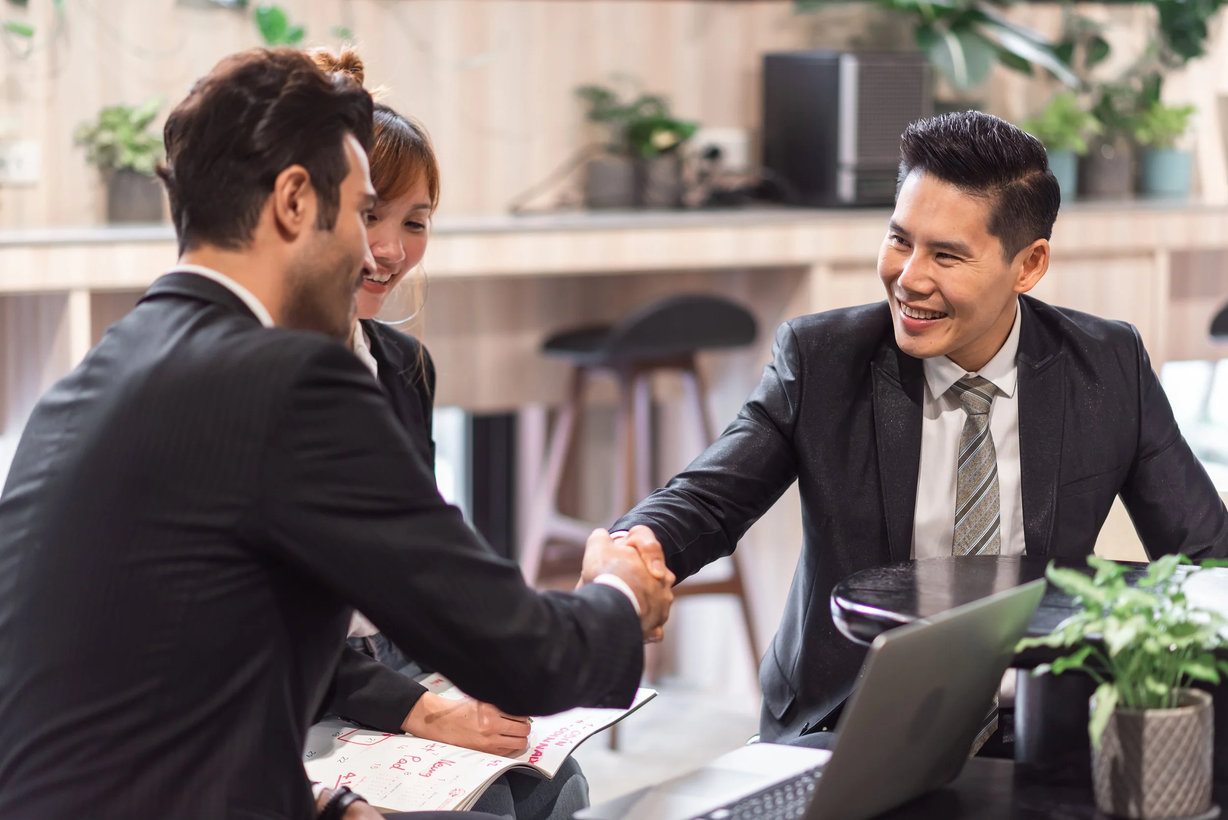 Three business professionals in formal attire shaking hands at a meeting table, smiling. One man is holding a notebook, and there is a laptop on the table. The background has potted plants and office equipment.