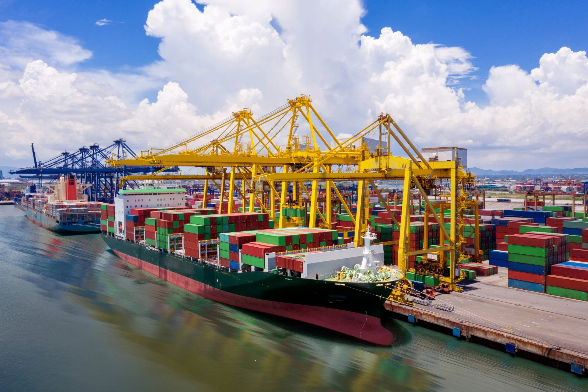 Large cargo ship carrying colorful shipping containers docked at a port with yellow and blue cranes under a partly cloudy sky.