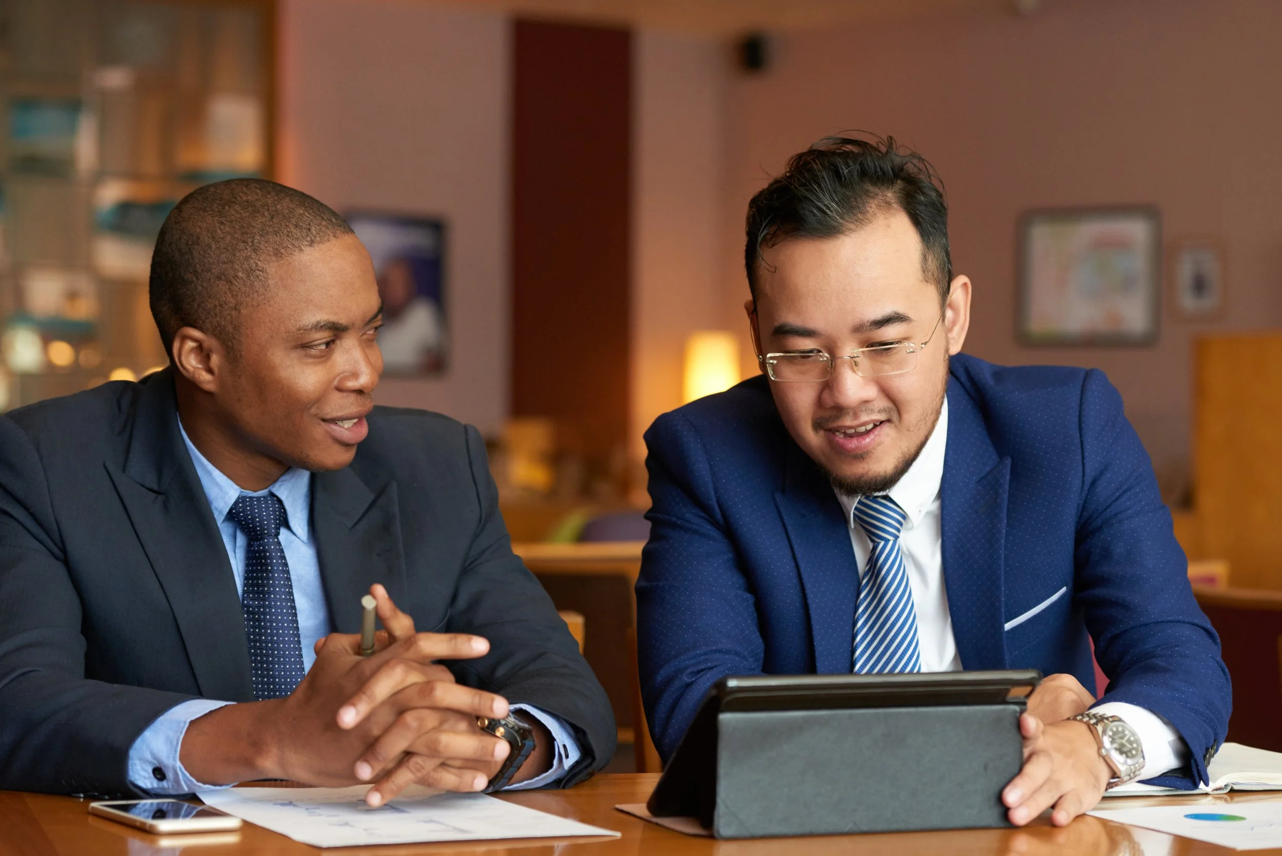 Two men in business suits sitting at a table, discussing and looking at a tablet computer in a professional office or conference room.