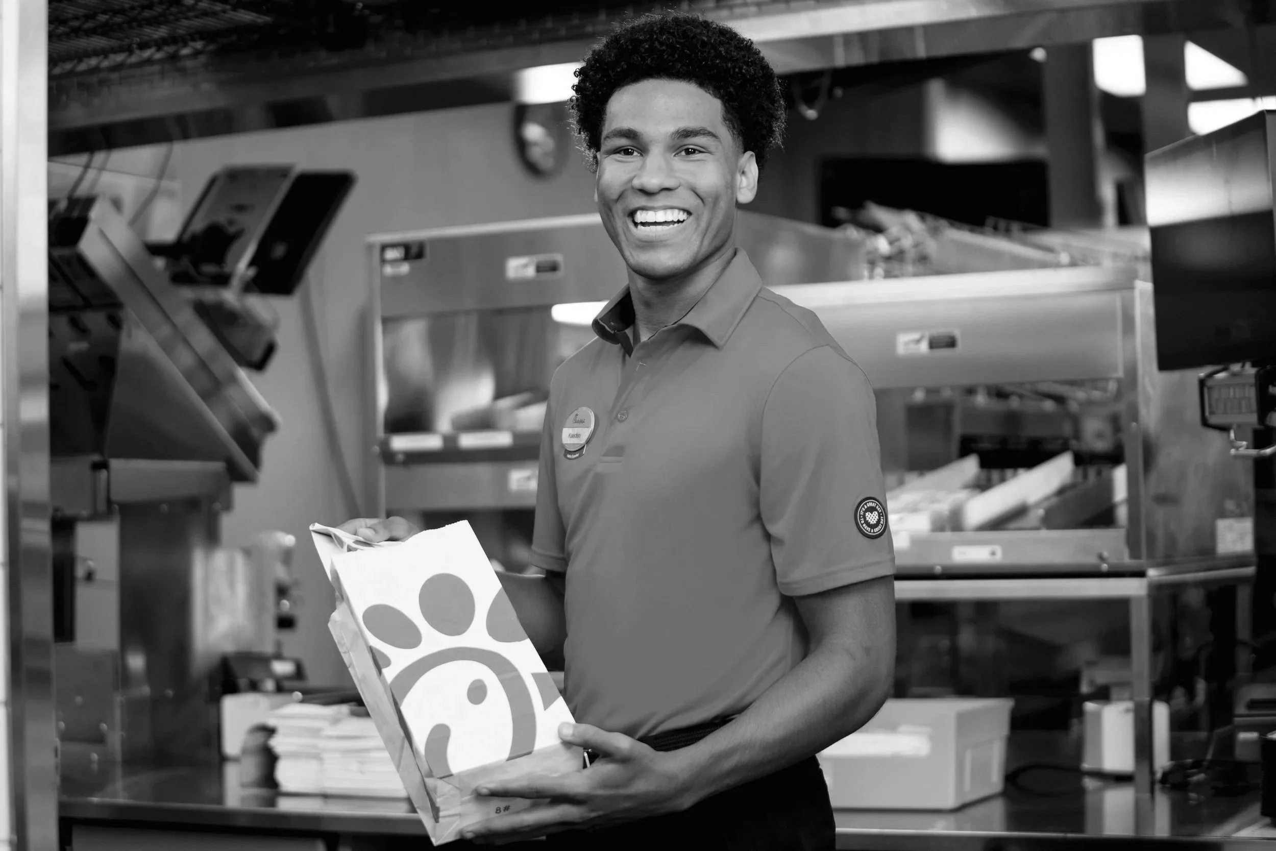 A young man with curly hair and a big smile, wearing a uniform, standing in an industrial setting, holding a Chick-fil-A food box.