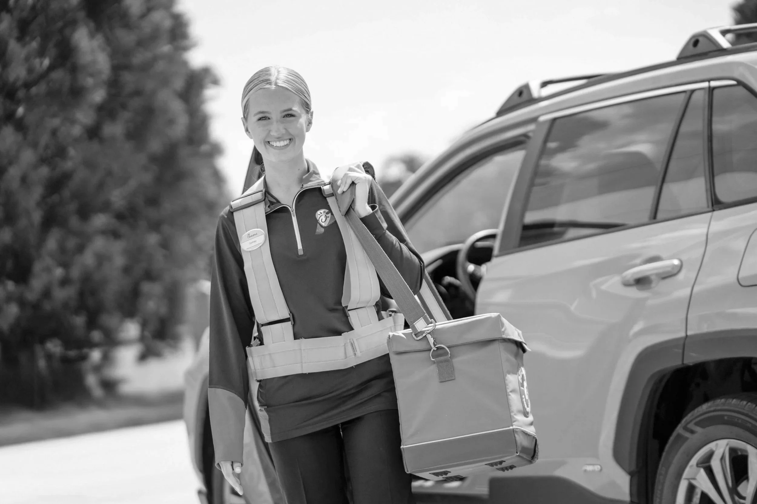 A smiling woman with blonde hair, wearing a sports jacket and carrying a backpack, stands by a parked SUV with a large delivery box in her hand.