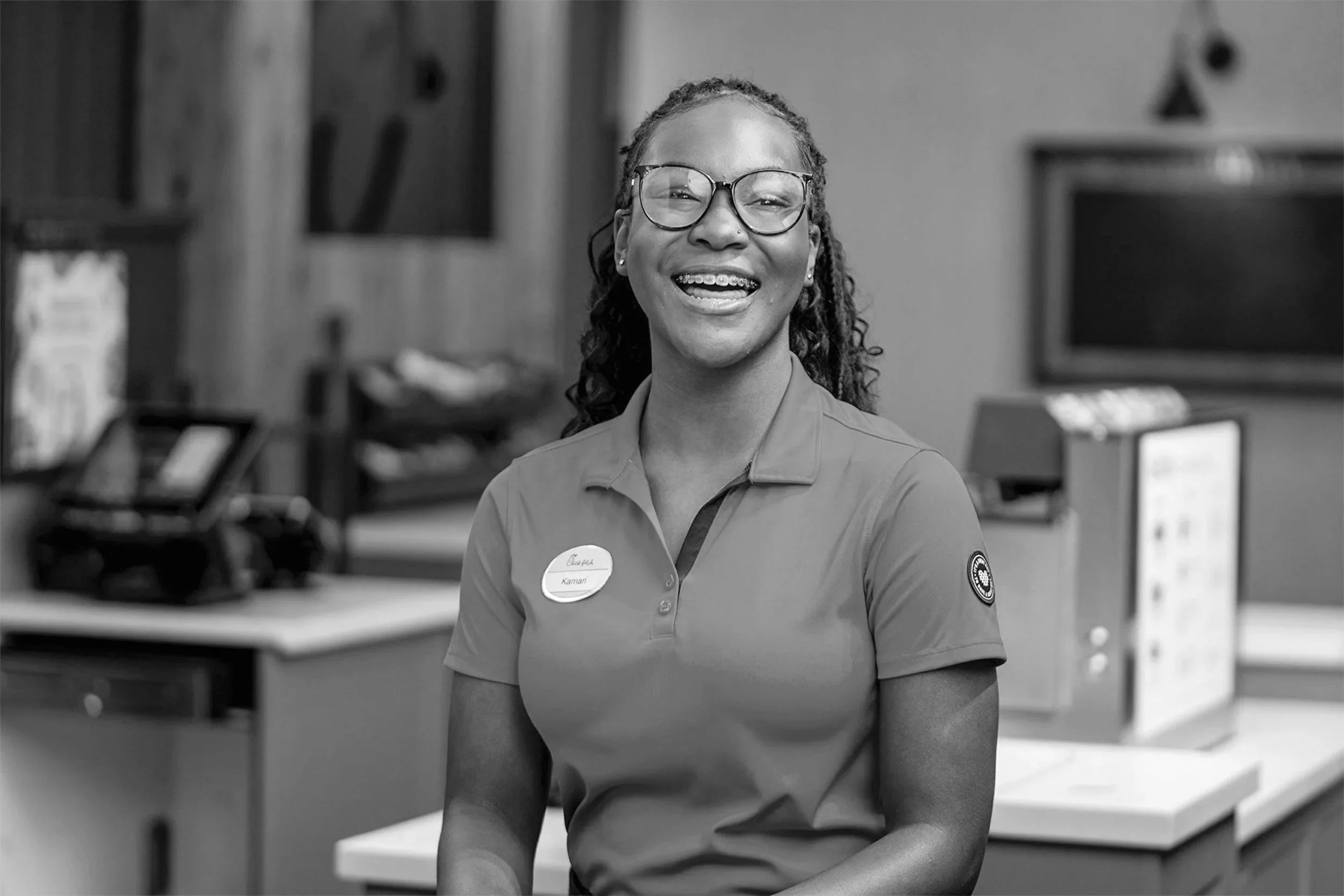 A woman with glasses, curly hair, and braces, smiling and wearing a uniform with a name tag in a retail or cafe setting.
