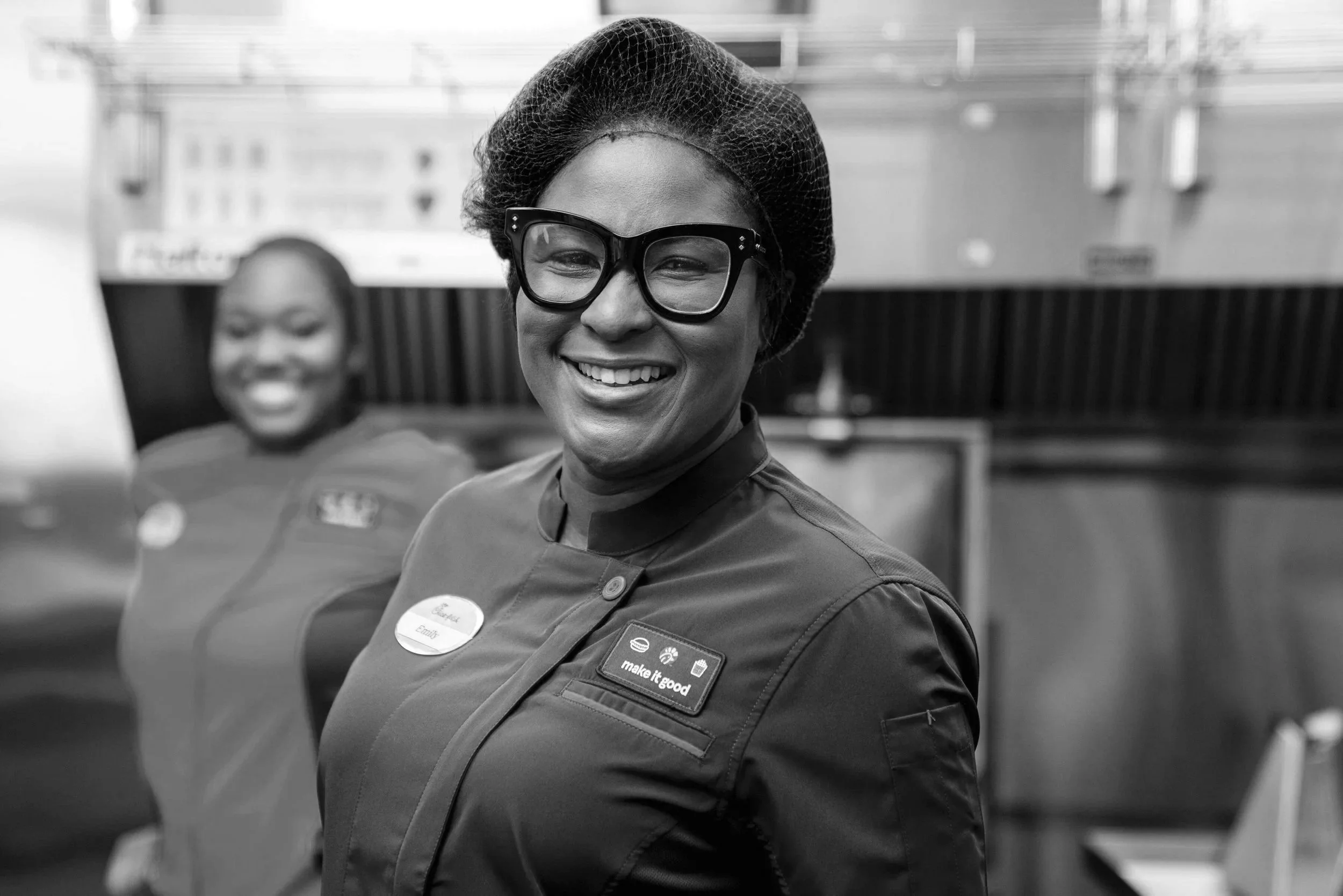Black and white photo of a smiling woman wearing glasses and a uniform with a name tag and a badge. She is in a work environment with another person in the background.