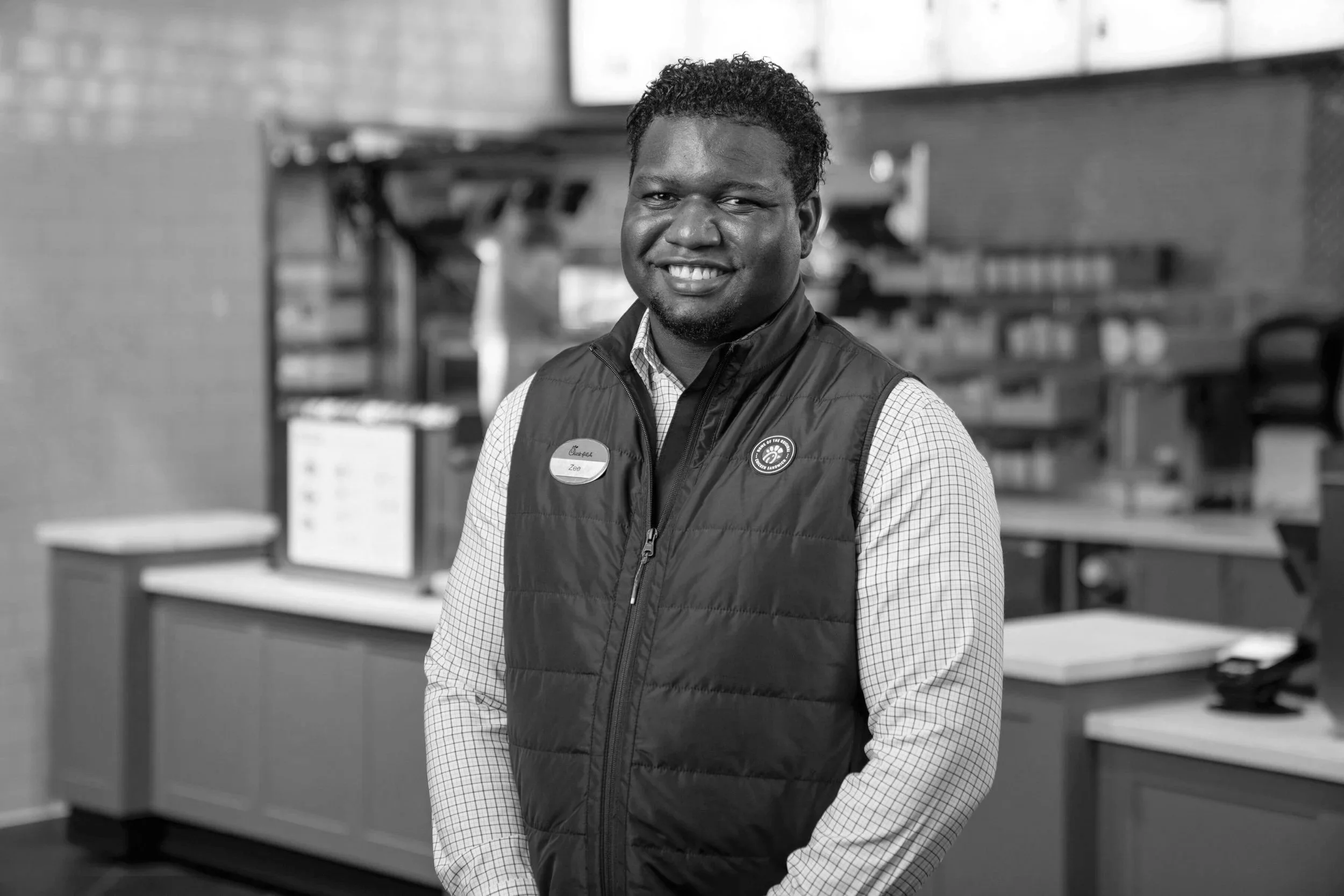 A man with curly hair wearing a checkered shirt and a vest, smiling for the camera in a fast-food restaurant kitchen.
