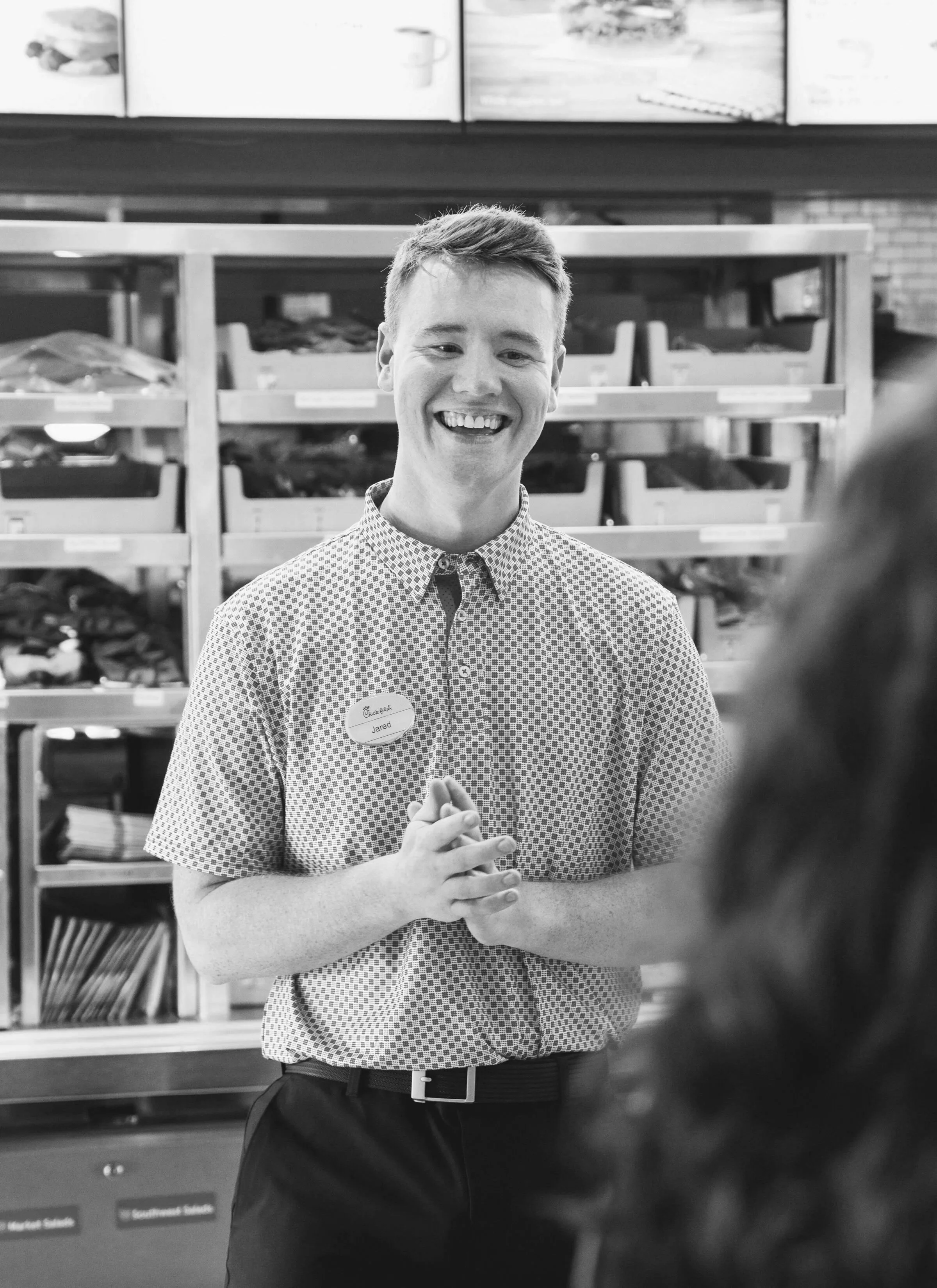 A smiling young man with short hair, wearing a checkered button-up shirt, standing in front of a shelving unit with labeled bins, possibly in a store or library. He has a name tag that says "Jared."