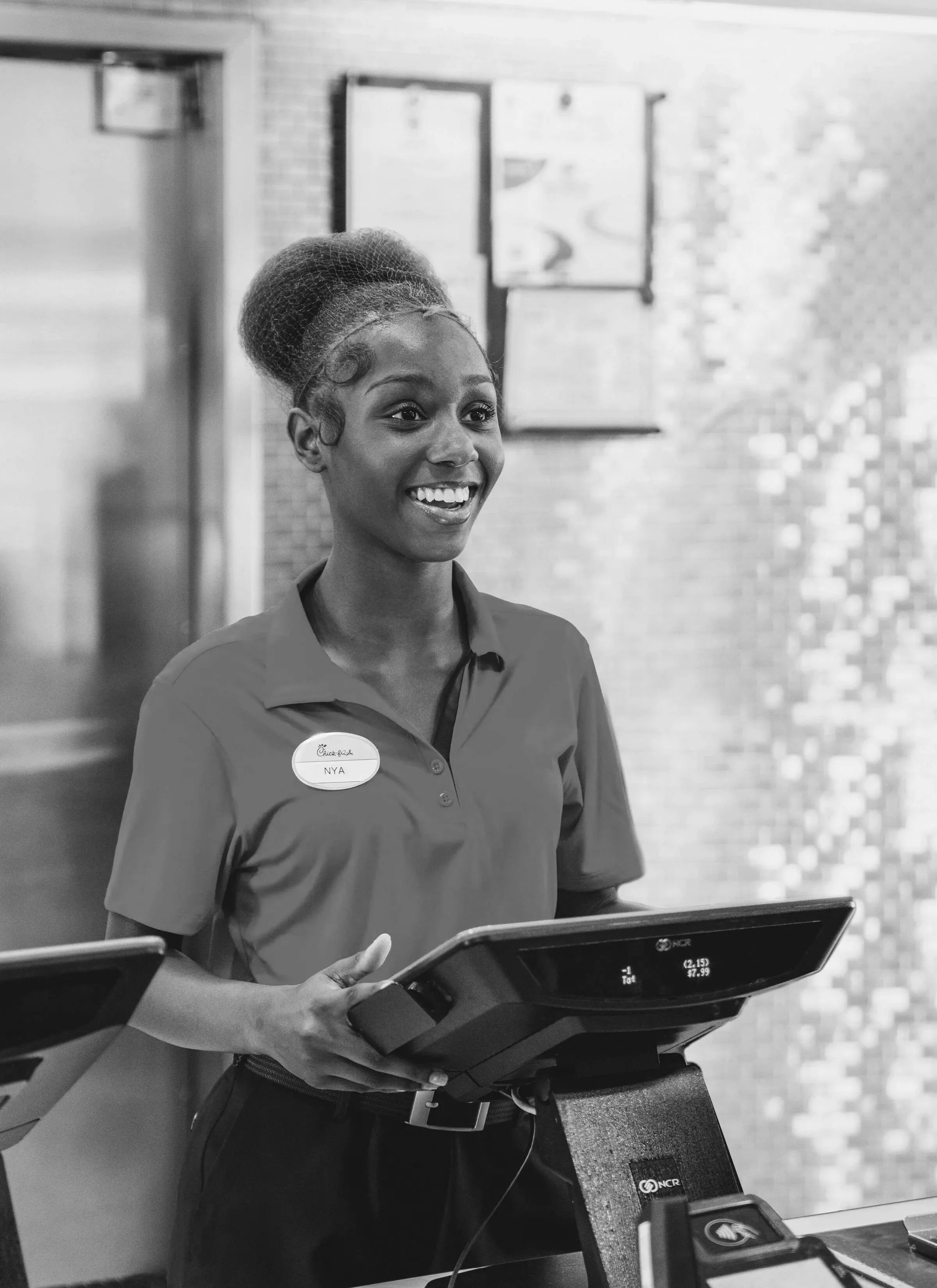 A young woman with dark skin and a natural hairstyle wearing a uniform with a name tag reading 'NYA' behind a payment terminal at a counter in a fast-food restaurant. She is smiling and looking to her left.