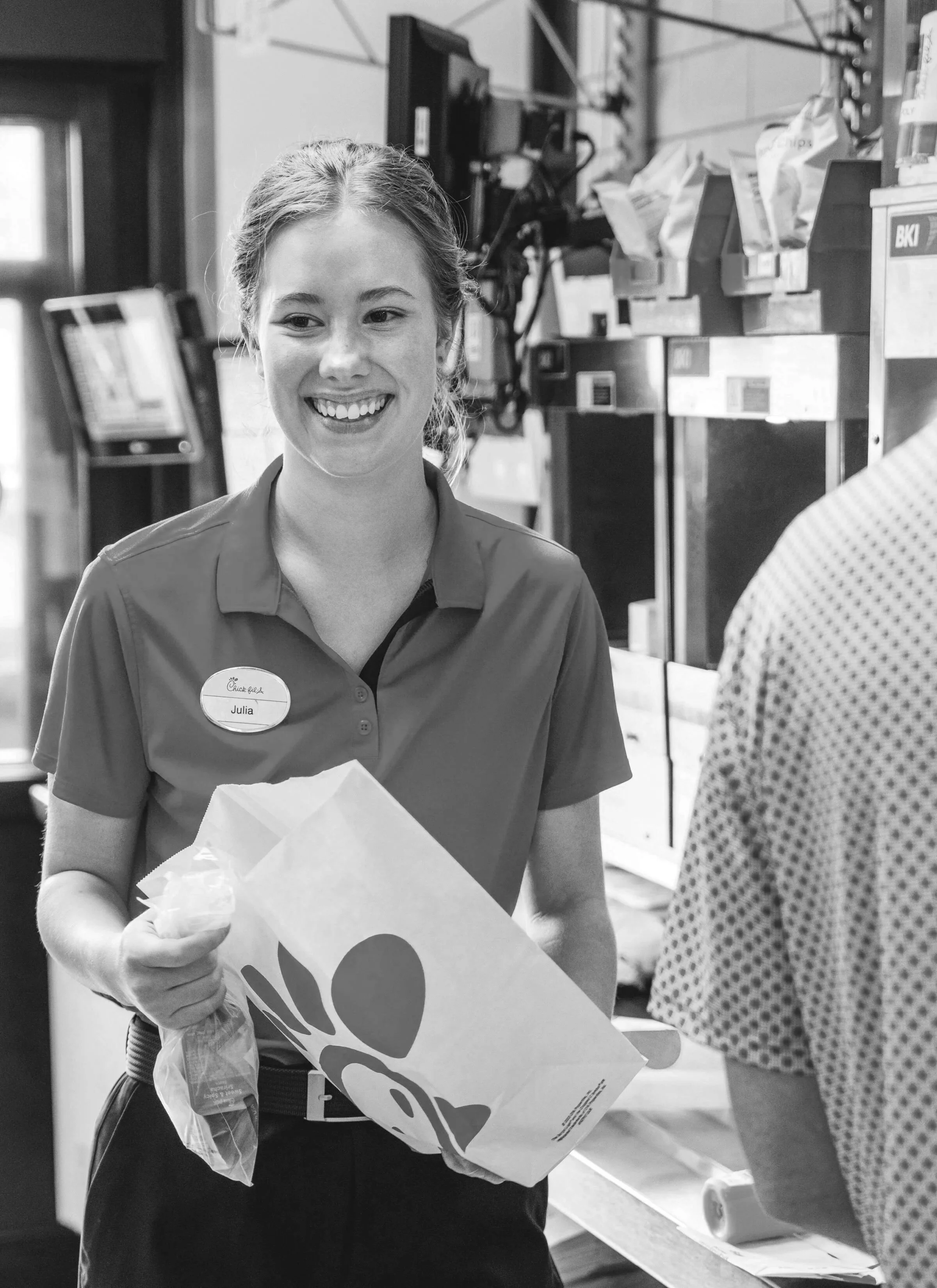 A young woman with a name tag that says Julia, smiling and holding a Chick-fil-A paper bag, working at a fast-food restaurant counter with snack attachments, and another person partially visible to the right, wearing patterned clothing.