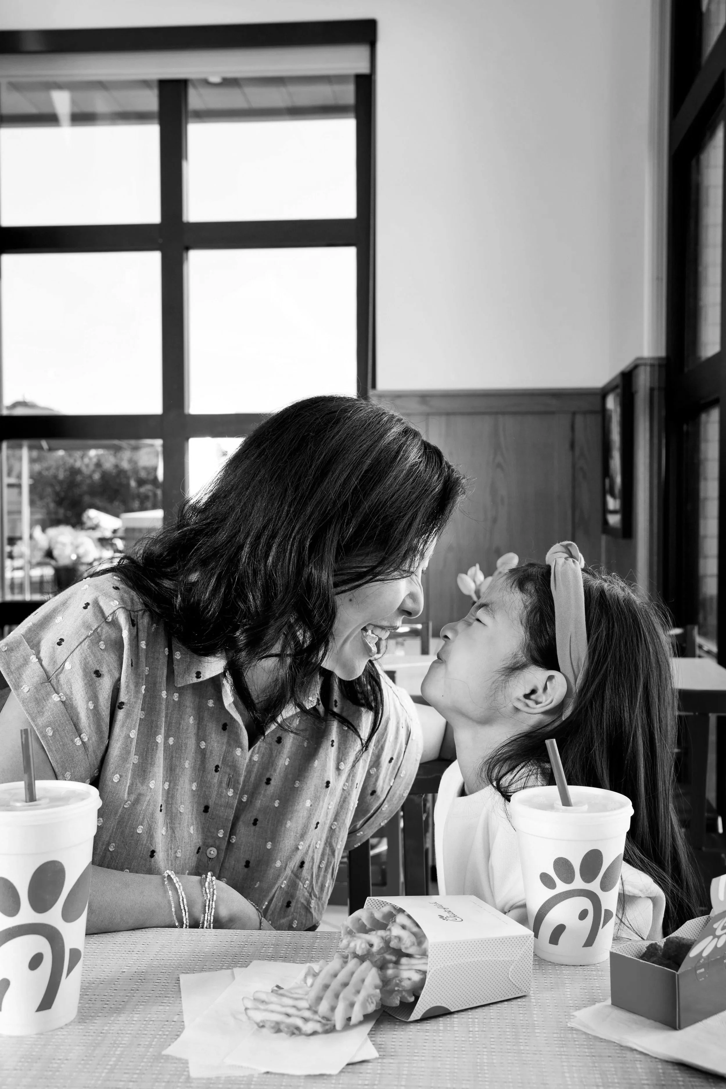 A woman and a young girl sitting at a fast food restaurant table, smiling and leaning close to each other. There are Chick-fil-A cups and food containers on the table.