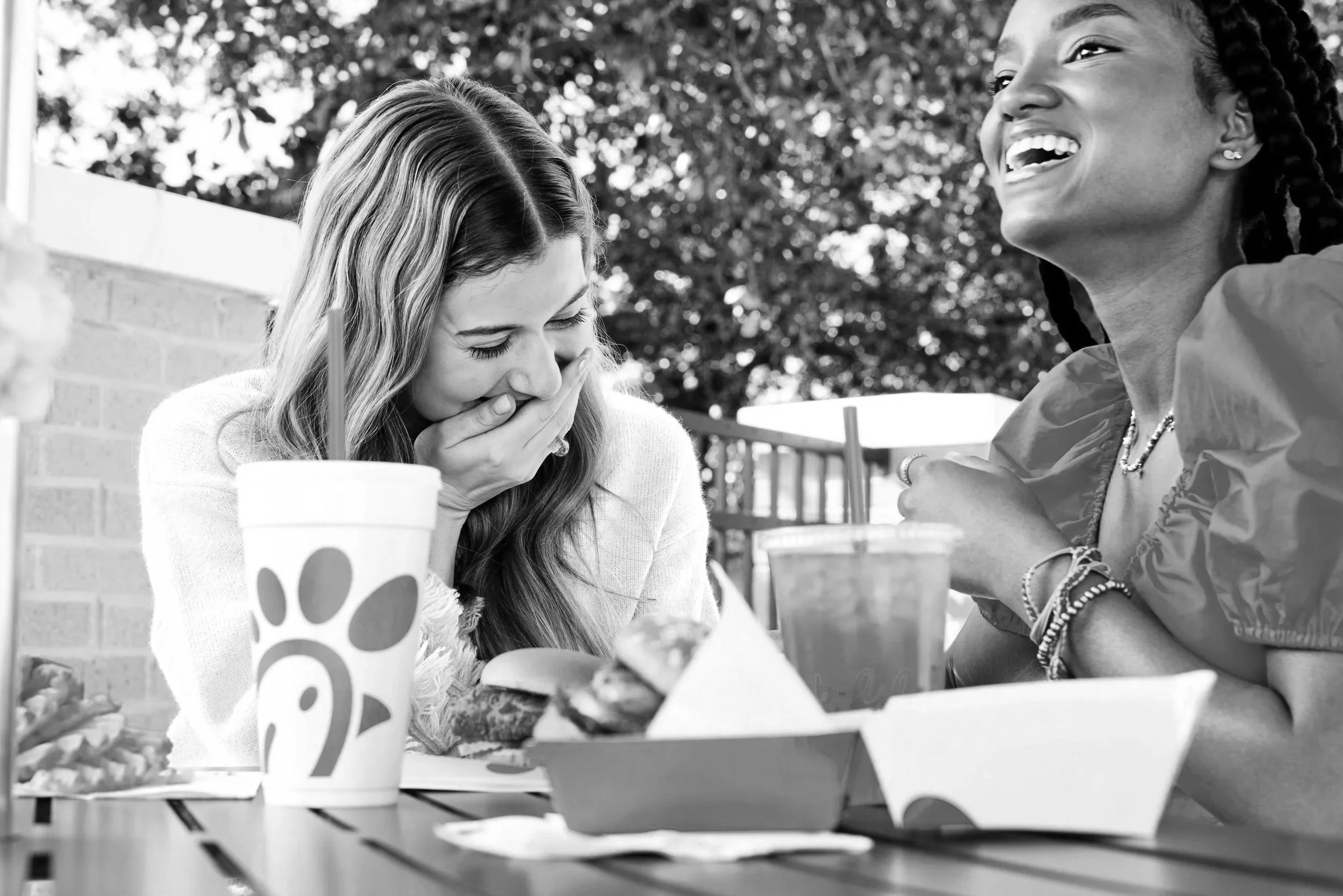 Two women sitting at a table outdoors, one is covering her mouth in surprise or laughter, and the other is smiling widely, wearing braided hair and jewelry. Food and drinks are on the table.