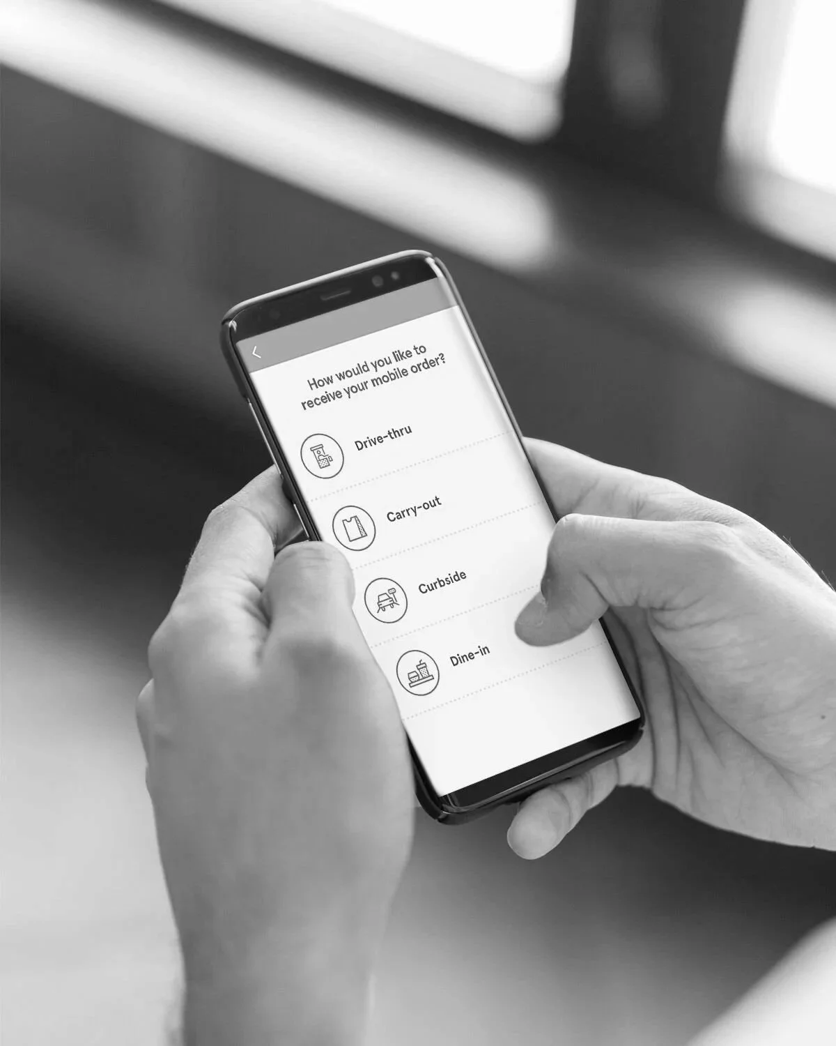 A person holding a smartphone displaying a menu with options for receiving a mobile order: Drive-thru, Carry-out, Curbside, Dine-in.