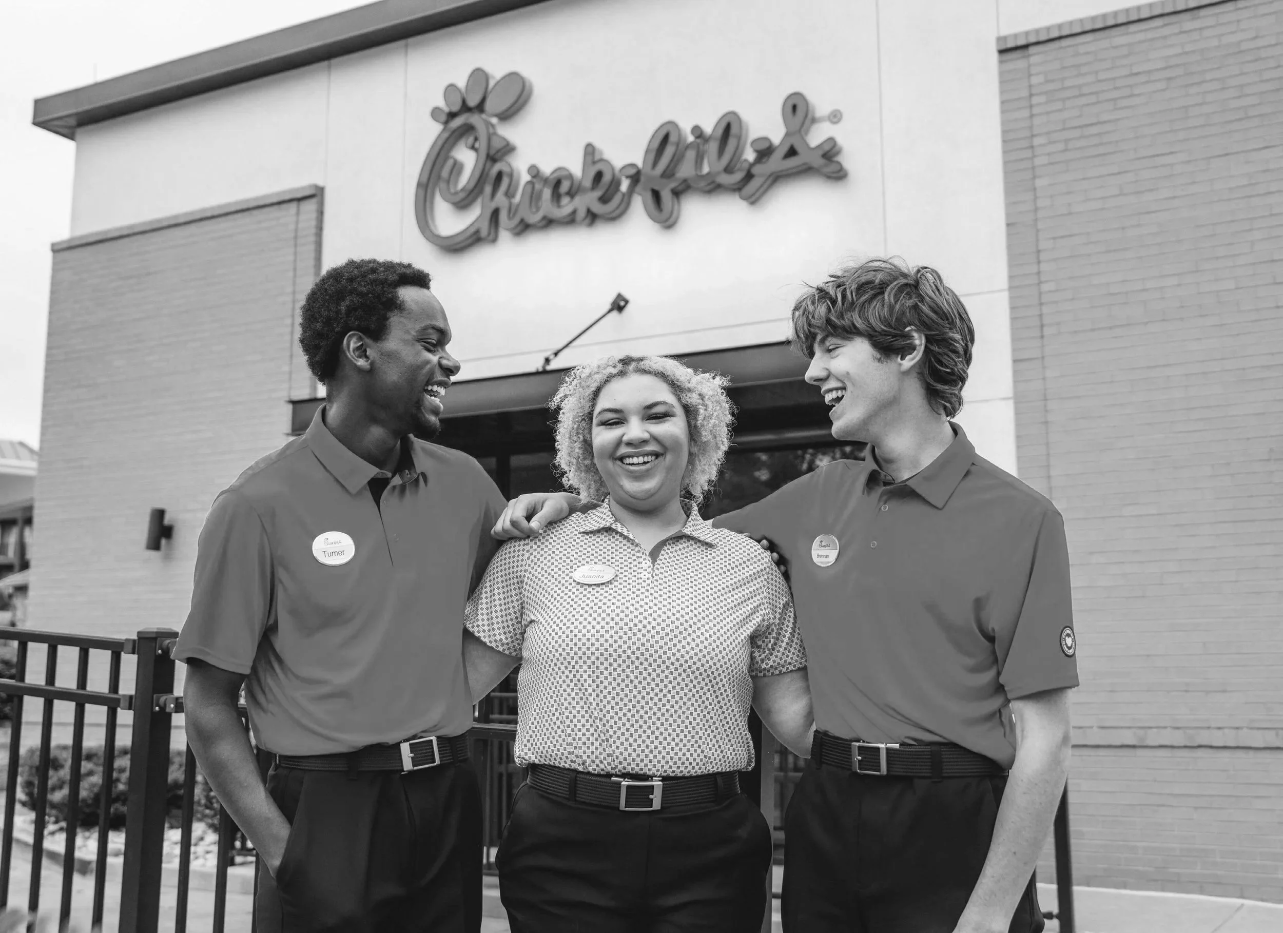 Three employees standing outside a Chick-fil-A restaurant, smiling and having a conversation. Two men are on either side of a woman in the middle, all wearing Chick-fil-A uniforms with name tags.