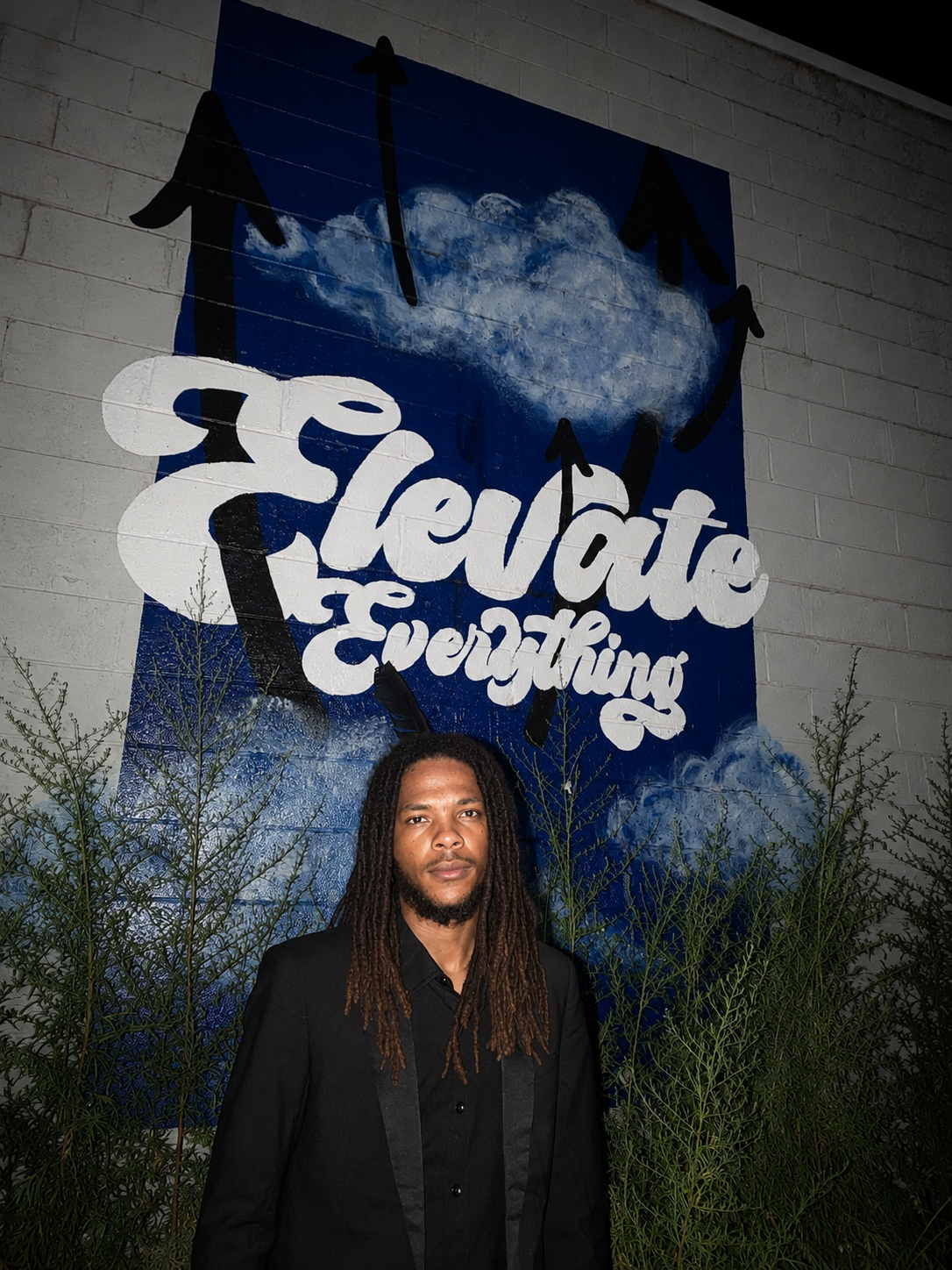 A man with long dreadlocks and a black suit standing in front of a mural that says 'Create everything' with a cloud and arrows pointing in different directions.