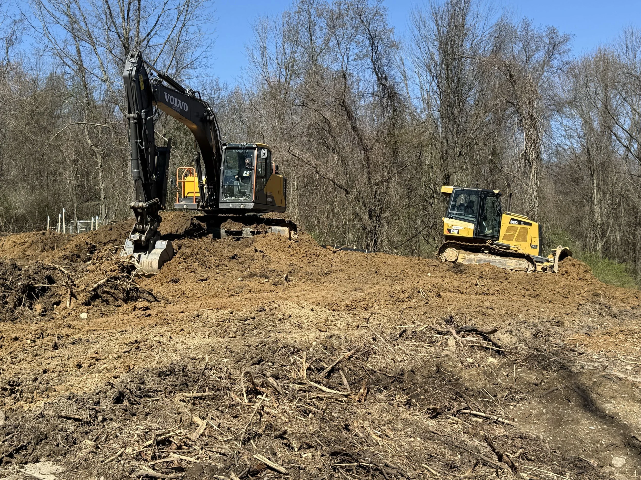 Construction site with two yellow and black excavators working on the ground, with a background of leafless trees and a clear blue sky.
