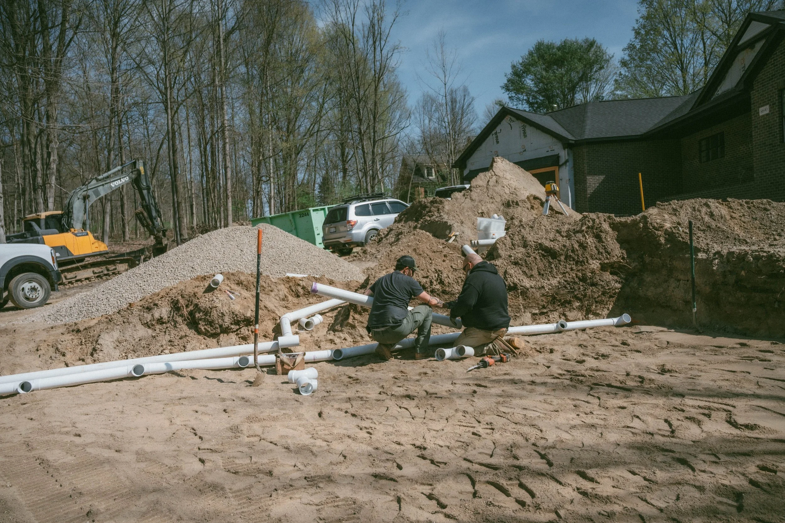 Construction workers installing underground pipes in a residential area, with a backhoe and parked cars nearby.