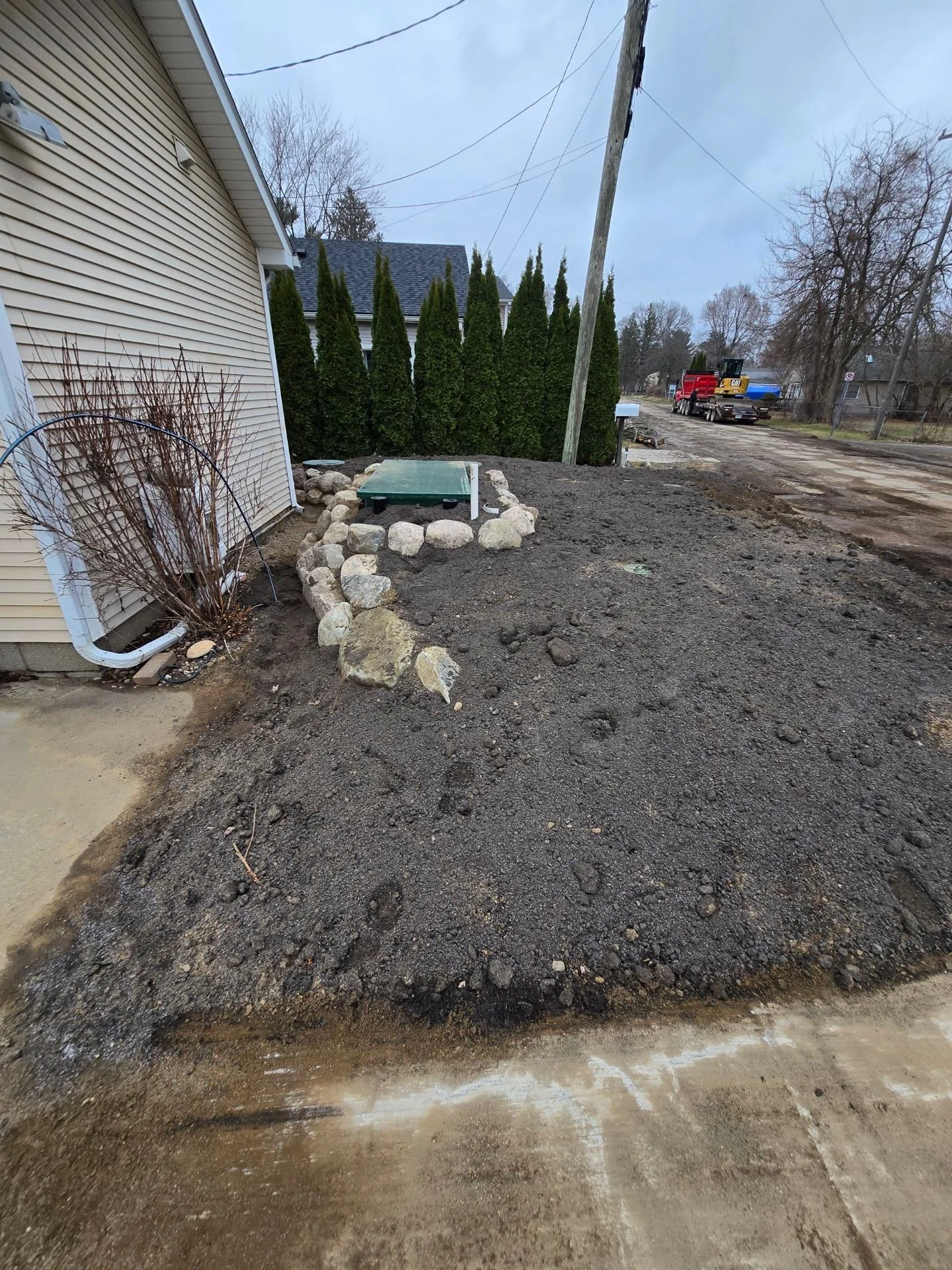 A backyard with dirt and gravel being prepared for landscaping or construction, with a row of trimmed bushes, a house, utility pole, and a distant construction vehicle on a cloudy day.