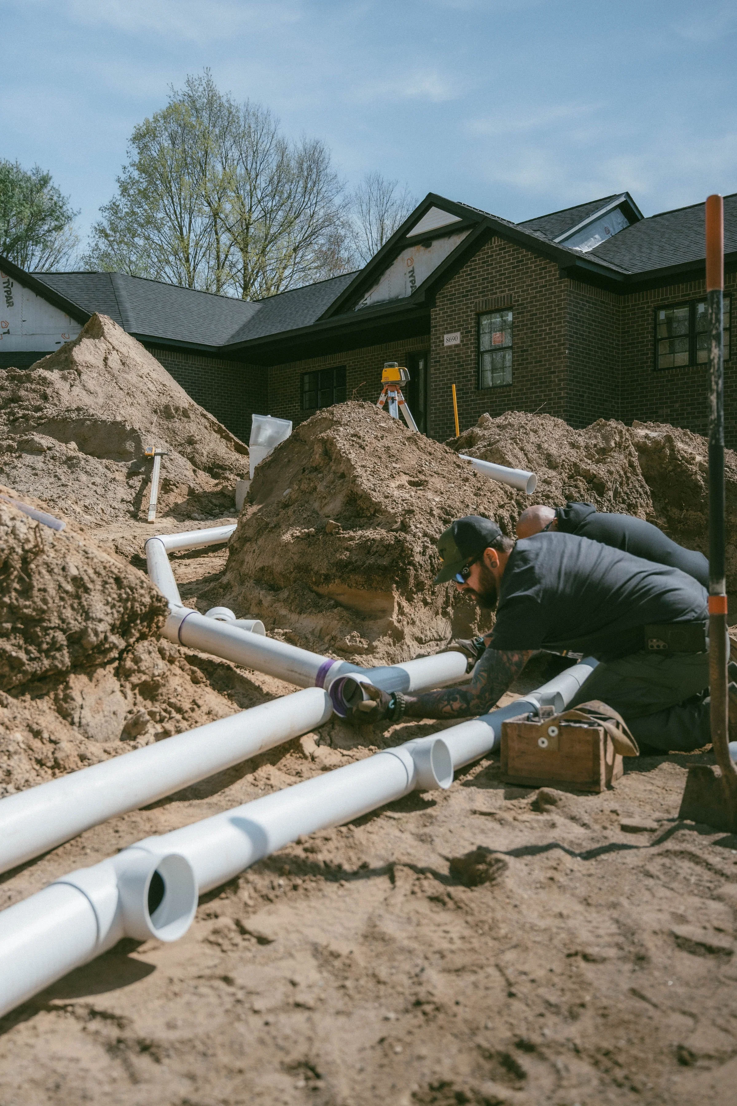 Janette & Sons workers installing underground white PVC pipes in the dirt in front of a brick house.