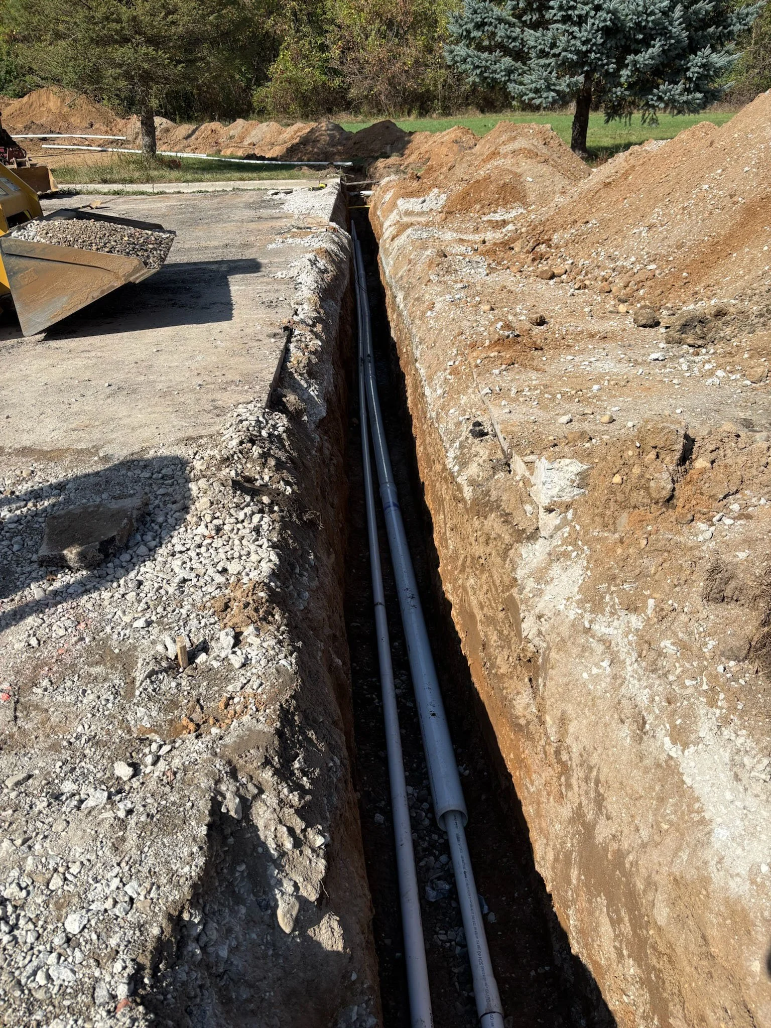 Construction site with a deep trench where two pipelines are being laid underground, surrounded by dirt and gravel, with trees in the background.