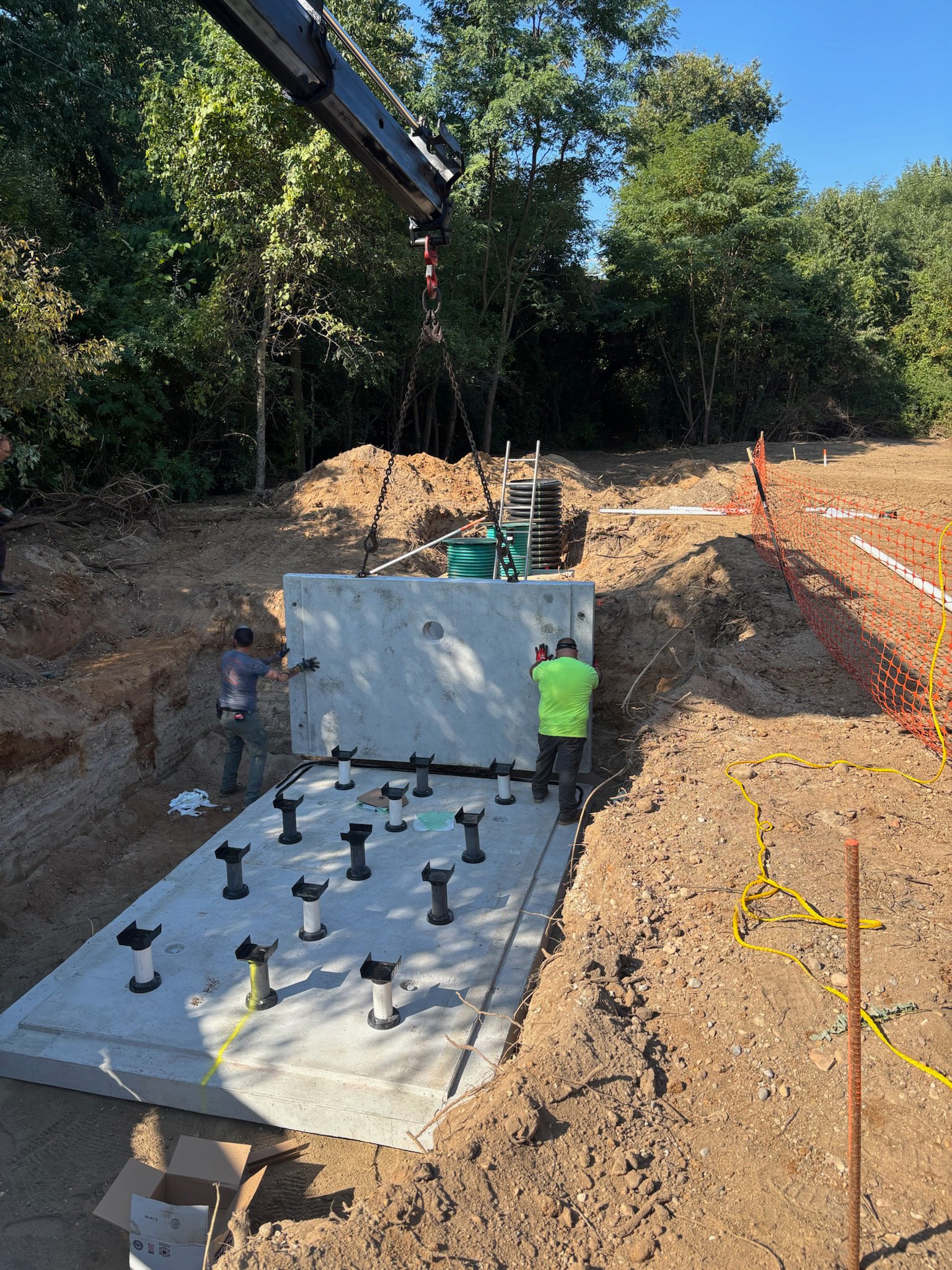 Workers install a large concrete utility vault with multiple pipe connections at a construction site surrounded by dirt and trees.