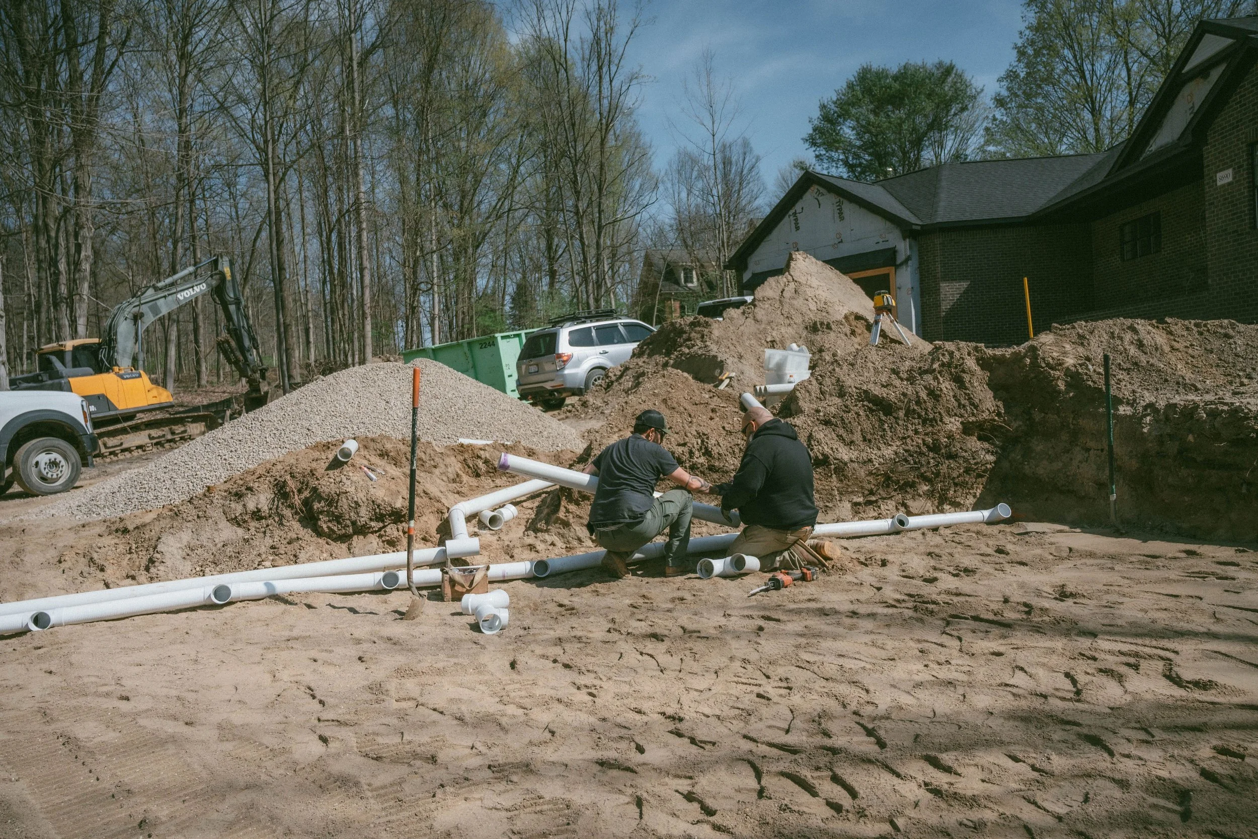 Construction workers installing white PVC pipes in a dug-up yard, with construction equipment and a partially built house in the background.