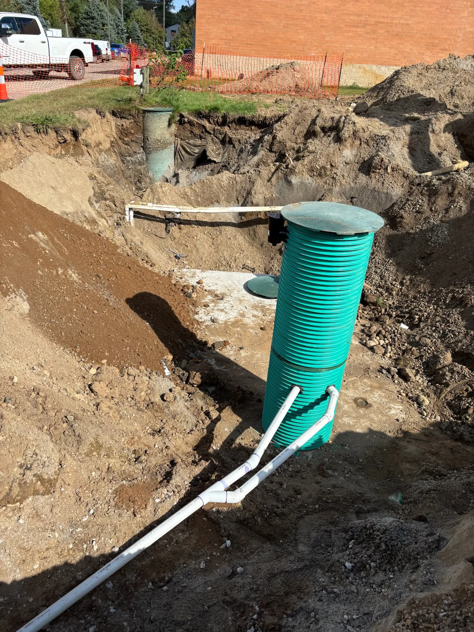 A construction site showing a large hole in the ground with exposed utility pipes, including a large green corrugated pipe, surrounded by dirt and gravel. Orange safety fencing is seen in the background near a brick building and parked vehicles.