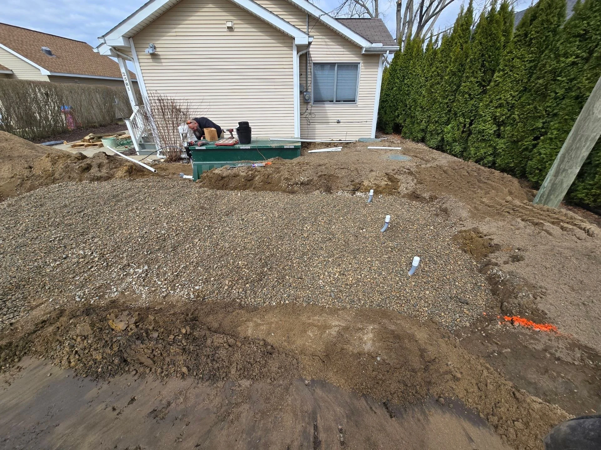 Backyard with dirt and gravel, indicating a landscaping or construction project, in front of a house with beige siding, a window with blinds, and a hedge on the right side.