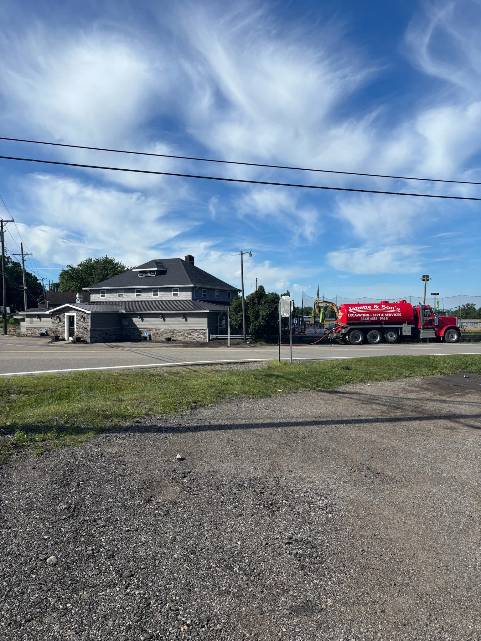 A street scene with a gravel foreground, a residential house with a dark roof and a sign on the roof, utility poles and wires, a red septic truck labeled 'Janette & Son's Excavating & Septic Services,' a lamppost, and a partly cloudy blue sky.