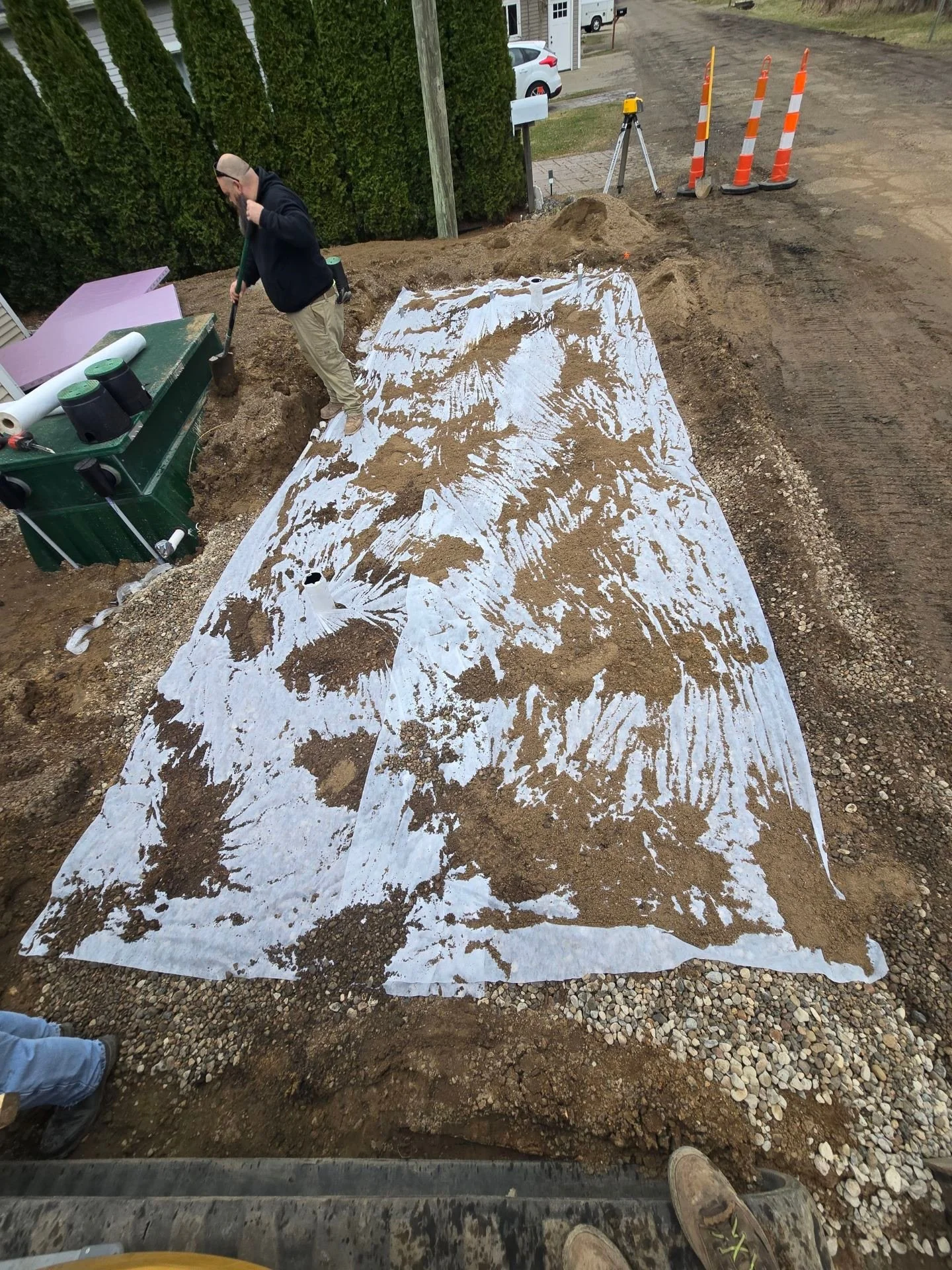 Construction worker shoveling soil or gravel on a construction site. The area is covered with a white tarp, and there are traffic cones, surveying equipment, and parked cars nearby.