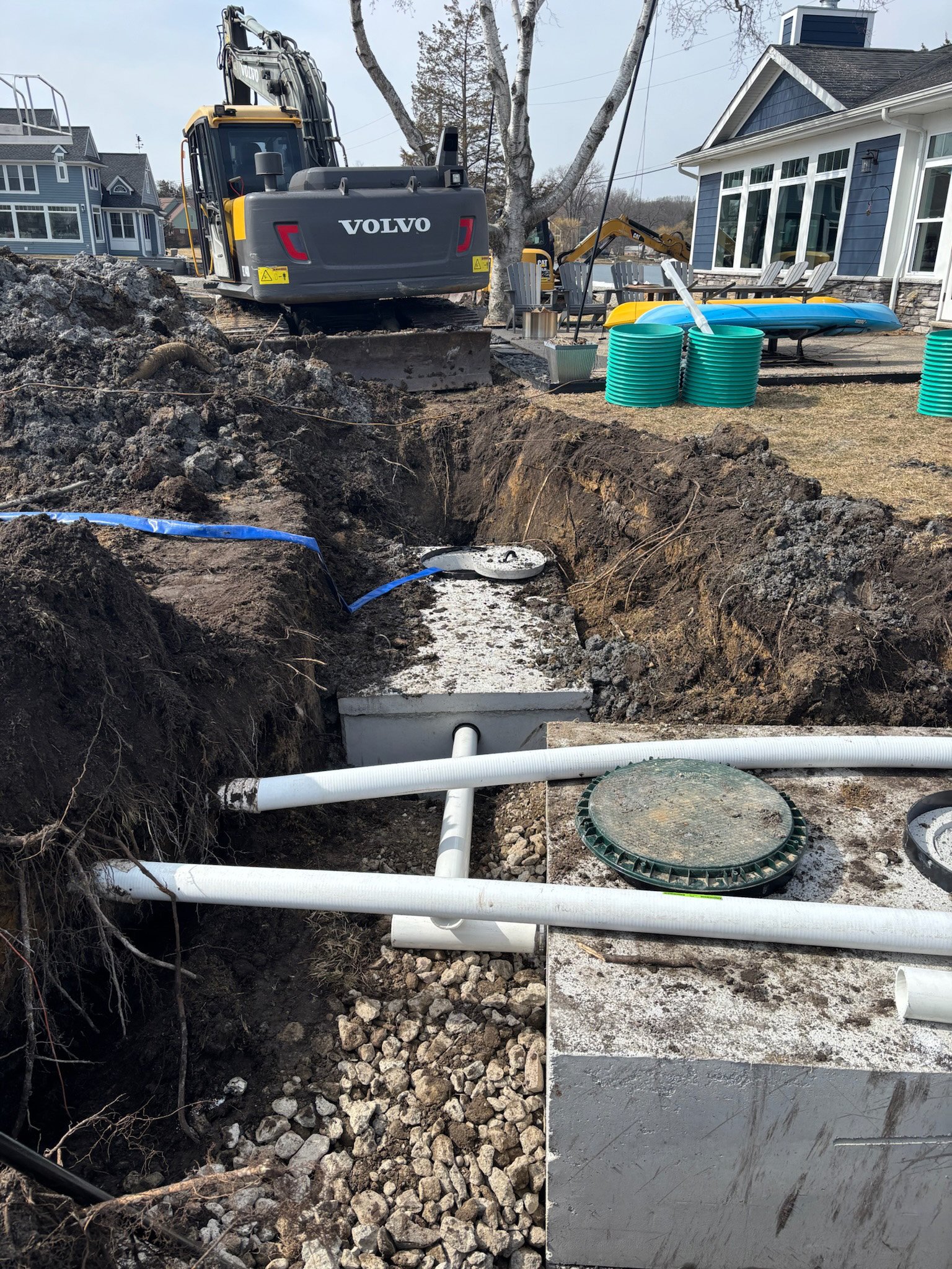 Construction site in a backyard where a trench is being dug for underground piping or utilities. A yellow and black excavator is positioned near a large hole in the ground. Several white pipes are placed across the trench, and green pipe fittings are nearby. In the background, there is a house with blue siding, outdoor patio furniture, and kayaks on the grass. A large tree stands next to the house.