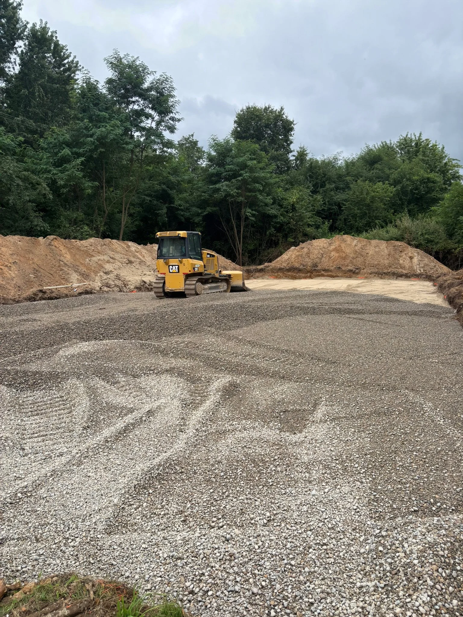 Construction site with a yellow Caterpillar bulldozer on a gravel-covered area, surrounded by dirt mounds and green trees under a cloudy sky.