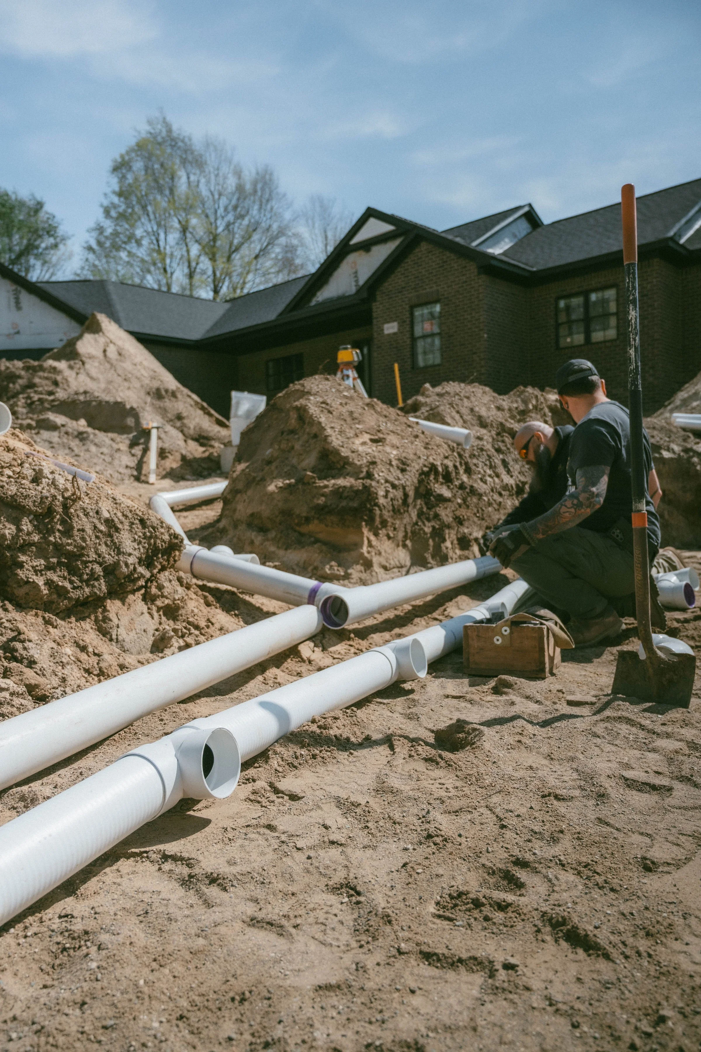 Workers are installing white PVC pipes in a dug trench in the yard of a house with a dark brick exterior. Piles of dirt are visible around the trench, and the workers are wearing safety glasses and gloves.