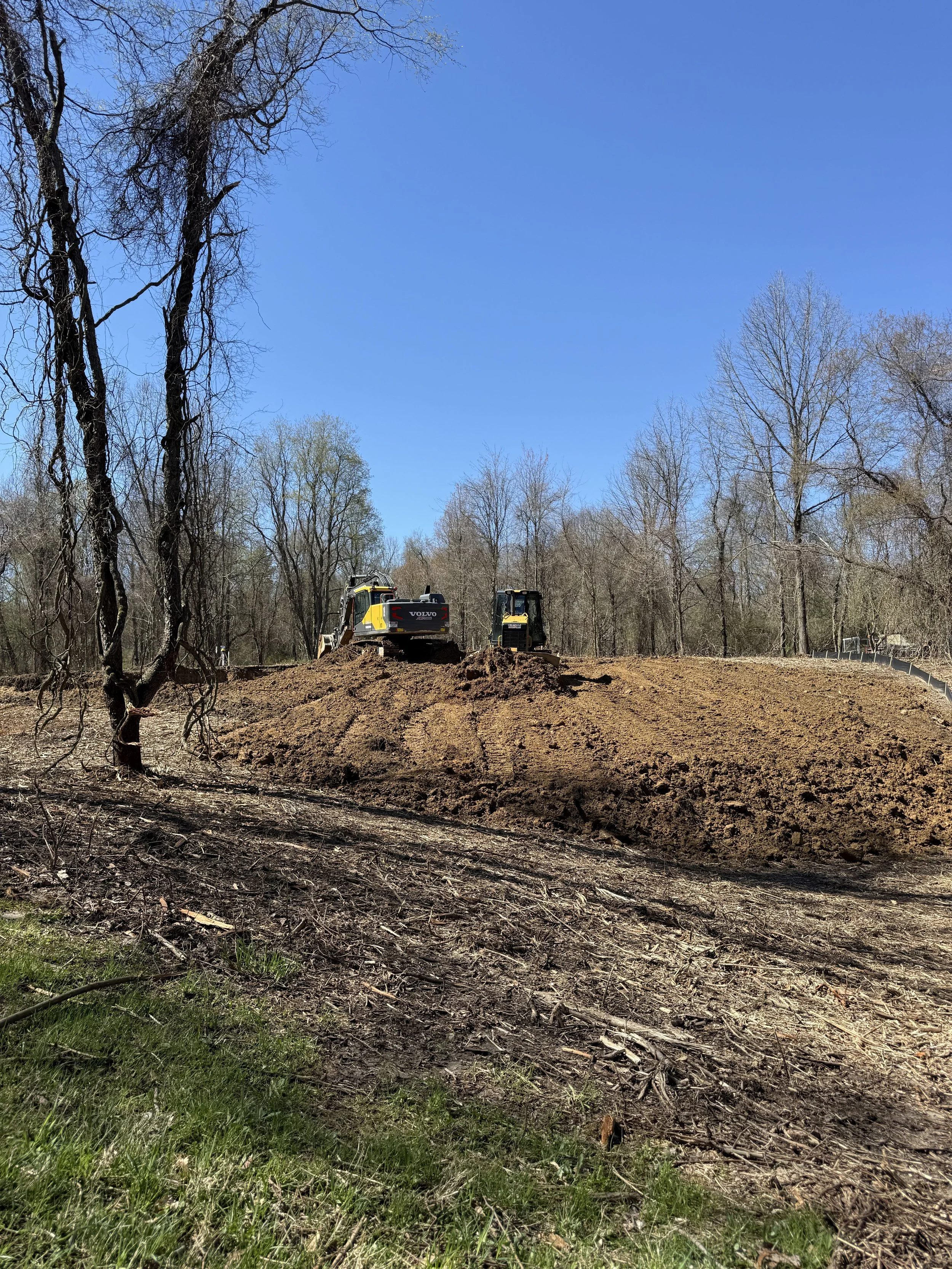 Two construction excavators working on a dirt clearing in a forested area with leafless trees under a blue sky.