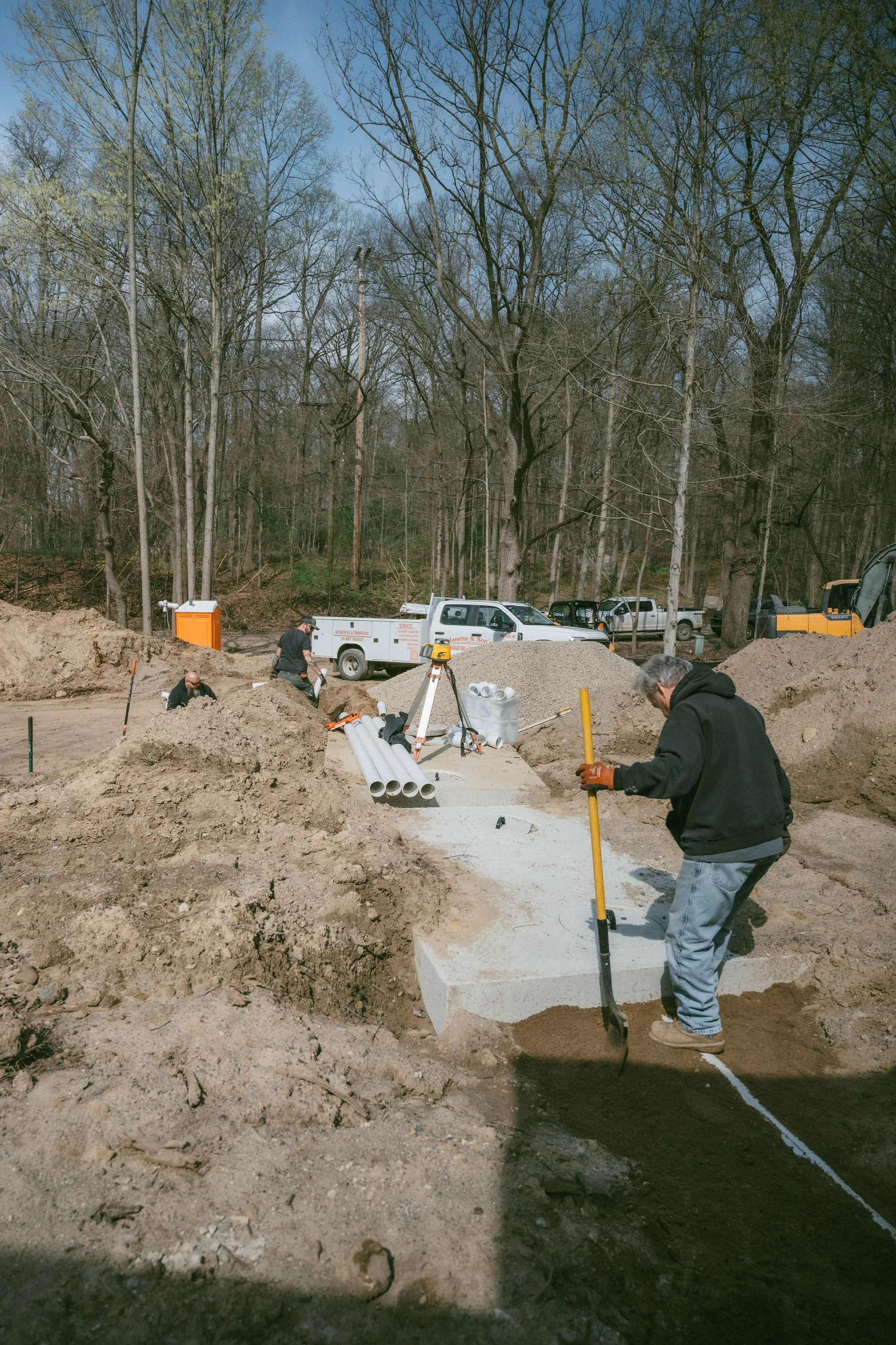 Workers laying concrete on a construction site with tools and equipment, surrounded by dirt and a wooded area in the background.