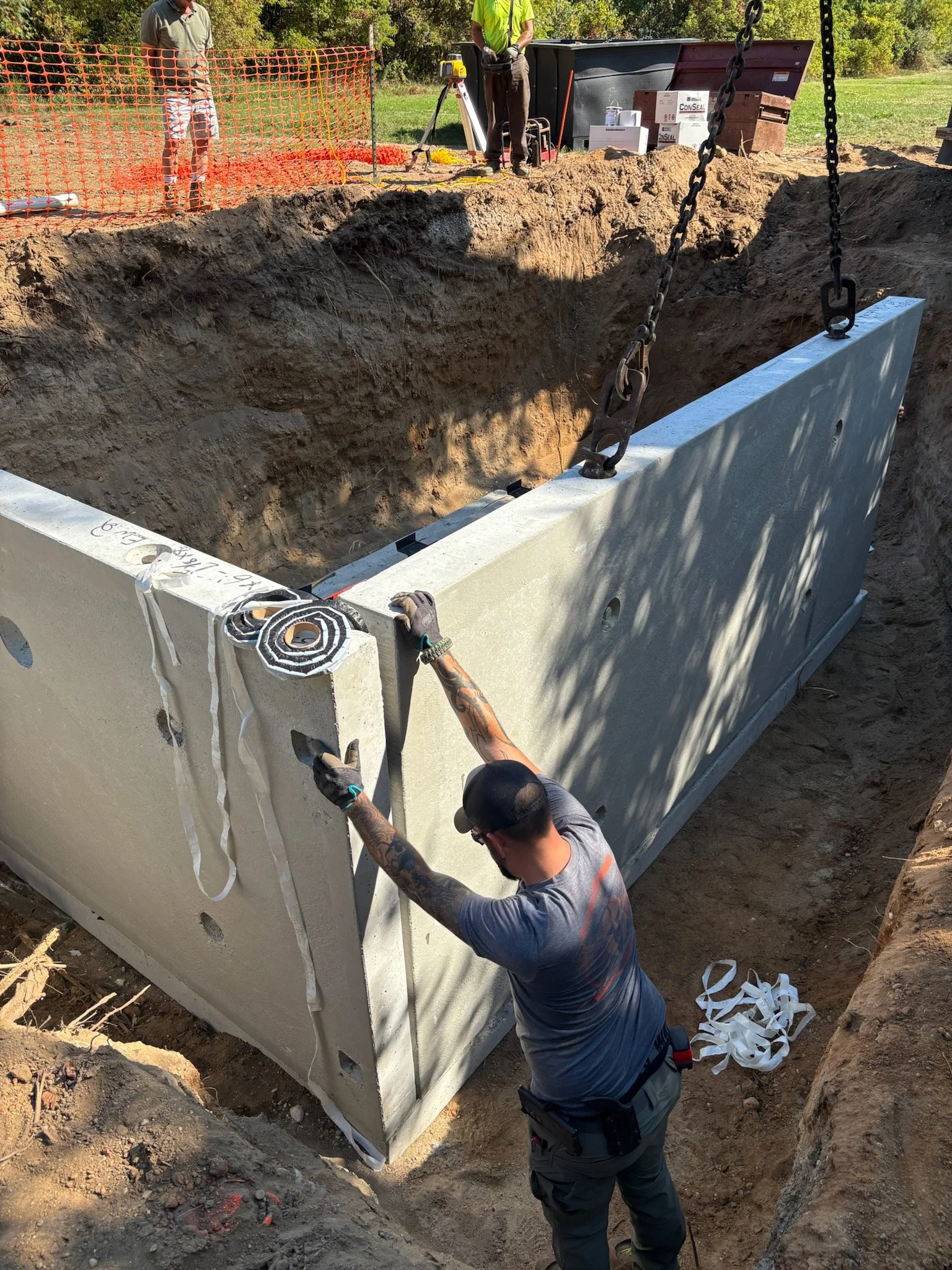 Construction workers installing a large concrete septic tank in an excavation site. One worker is holding the tank while another is overseeing the placement.