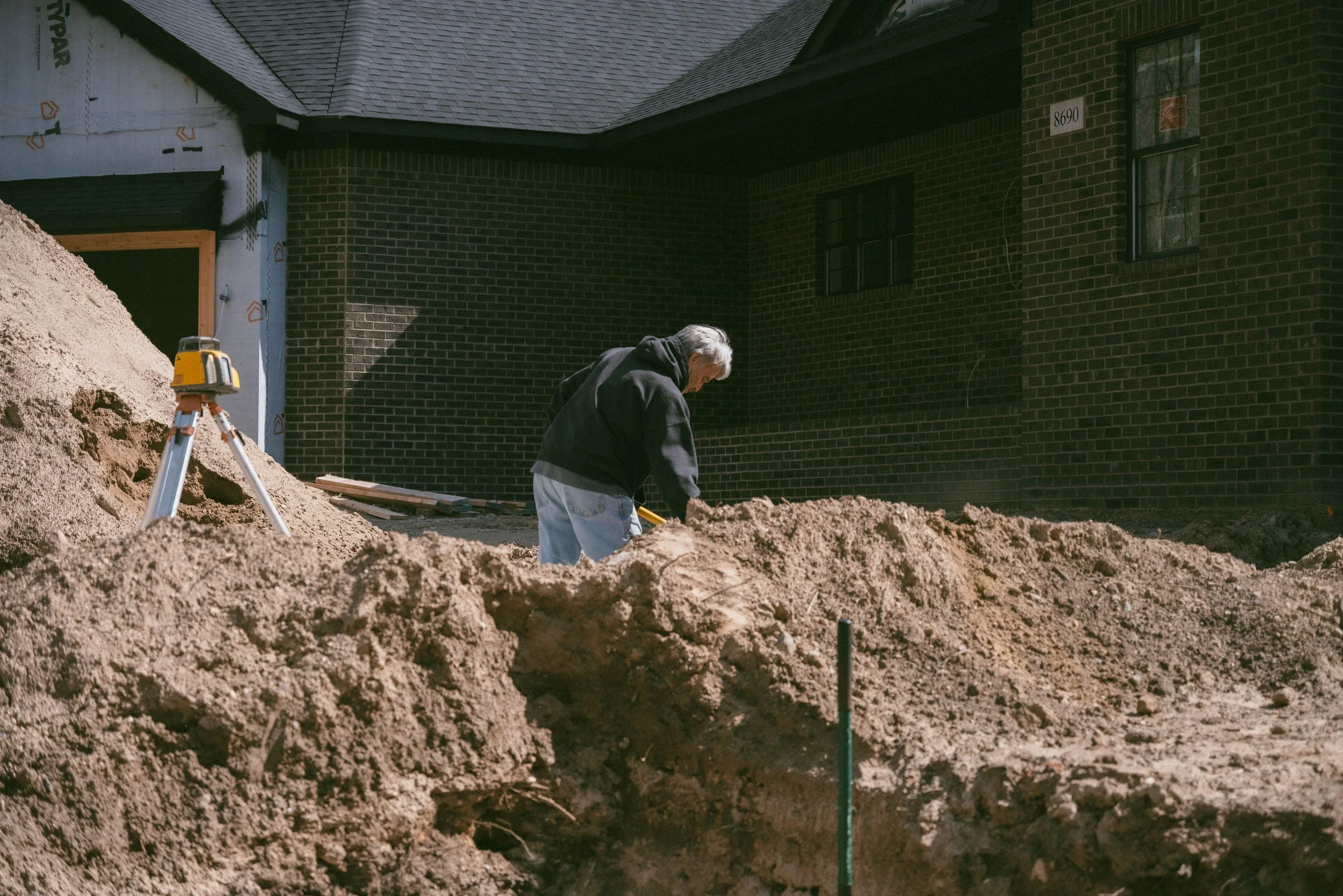 A person working on a construction site in front of a brick house, with a pile of dirt and construction tools nearby.