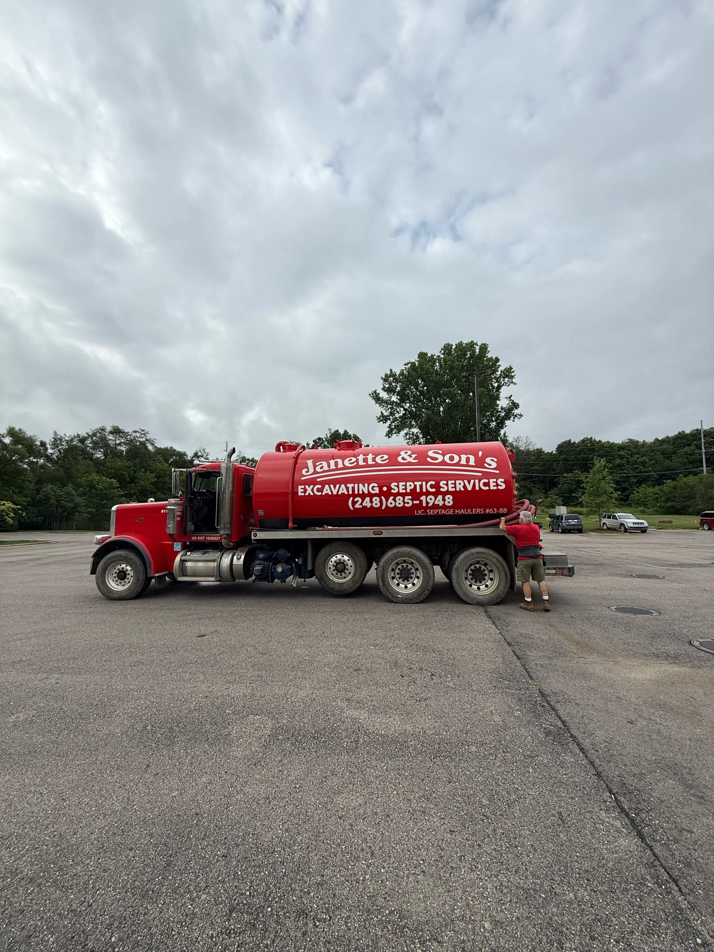 Red septic service truck parked in an empty parking lot with a man standing near it, in front of a large tree under a cloudy sky.