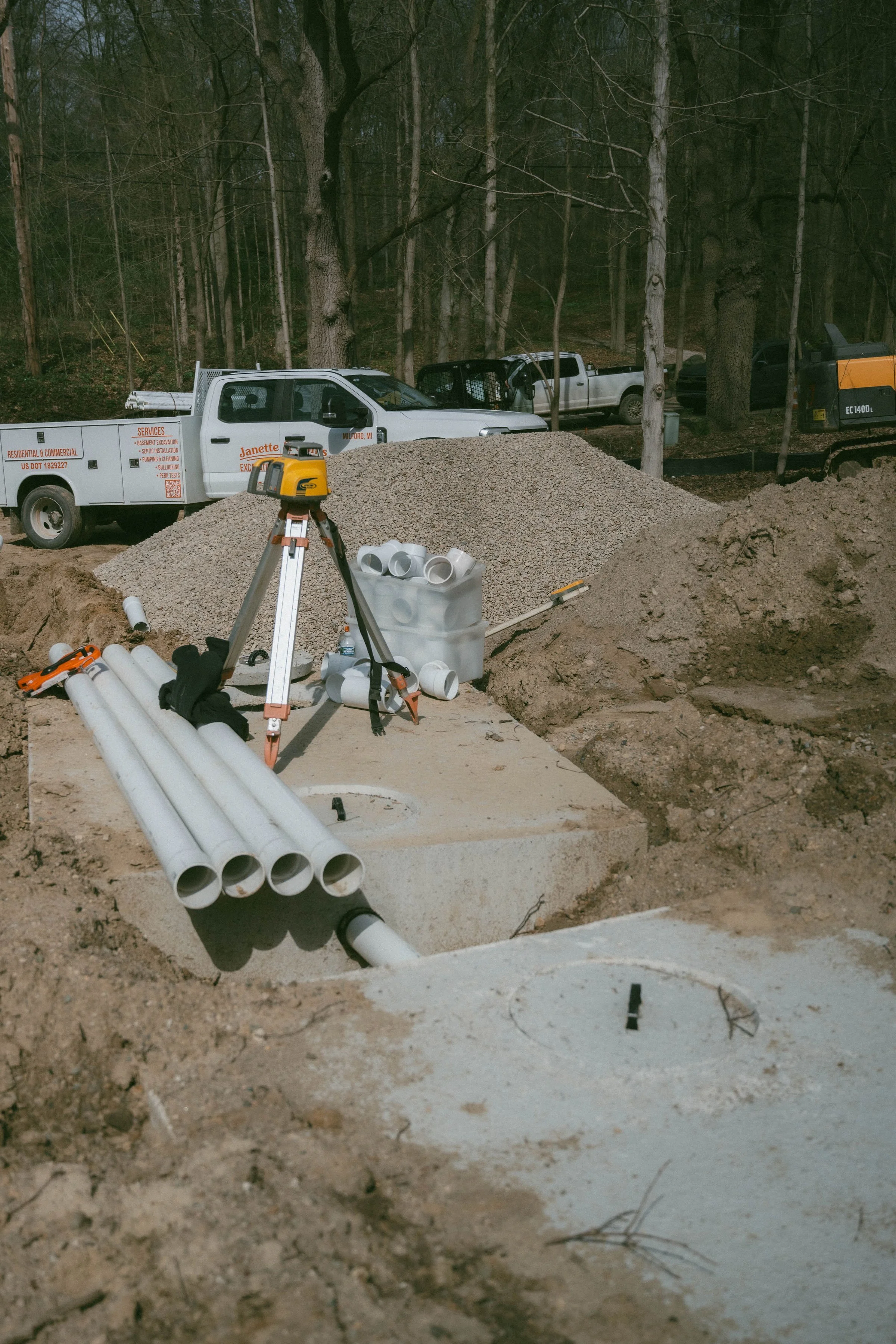 Construction site with underground piping, construction tools, gravel pile, pickup trucks, and an excavator in a wooded area.