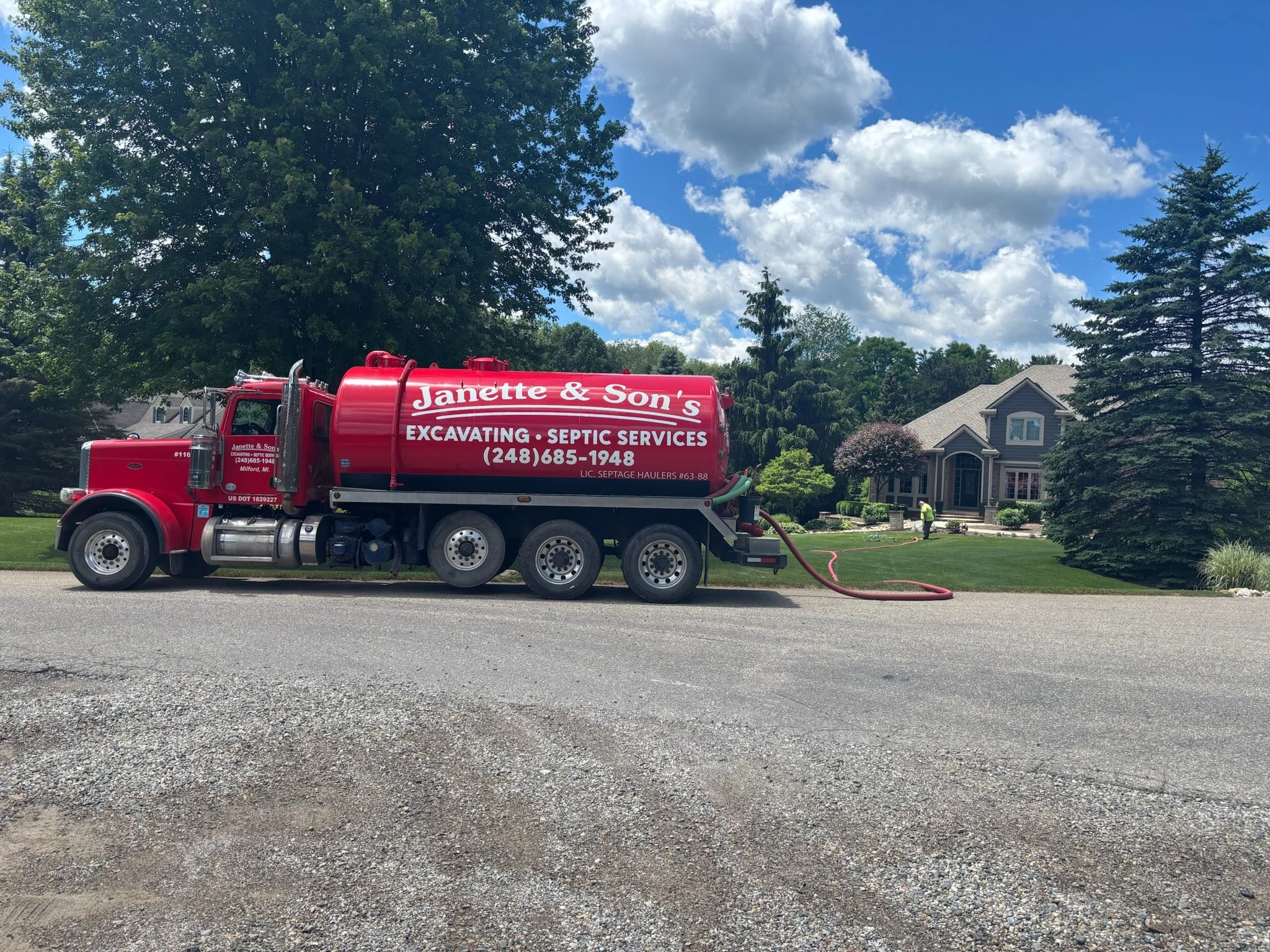 A red truck with white lettering advertising Janette & Son's Excavating and Septic Services parked on a residential street. The background shows a large house, trees, and a blue sky with clouds.
