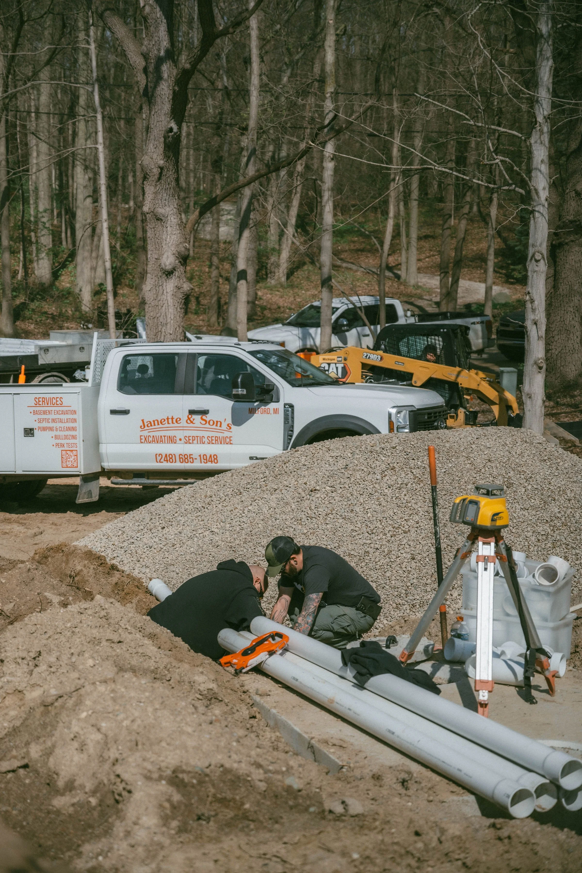 Construction workers assembling PVC pipes at a job site with trucks and equipment in a wooded area.
