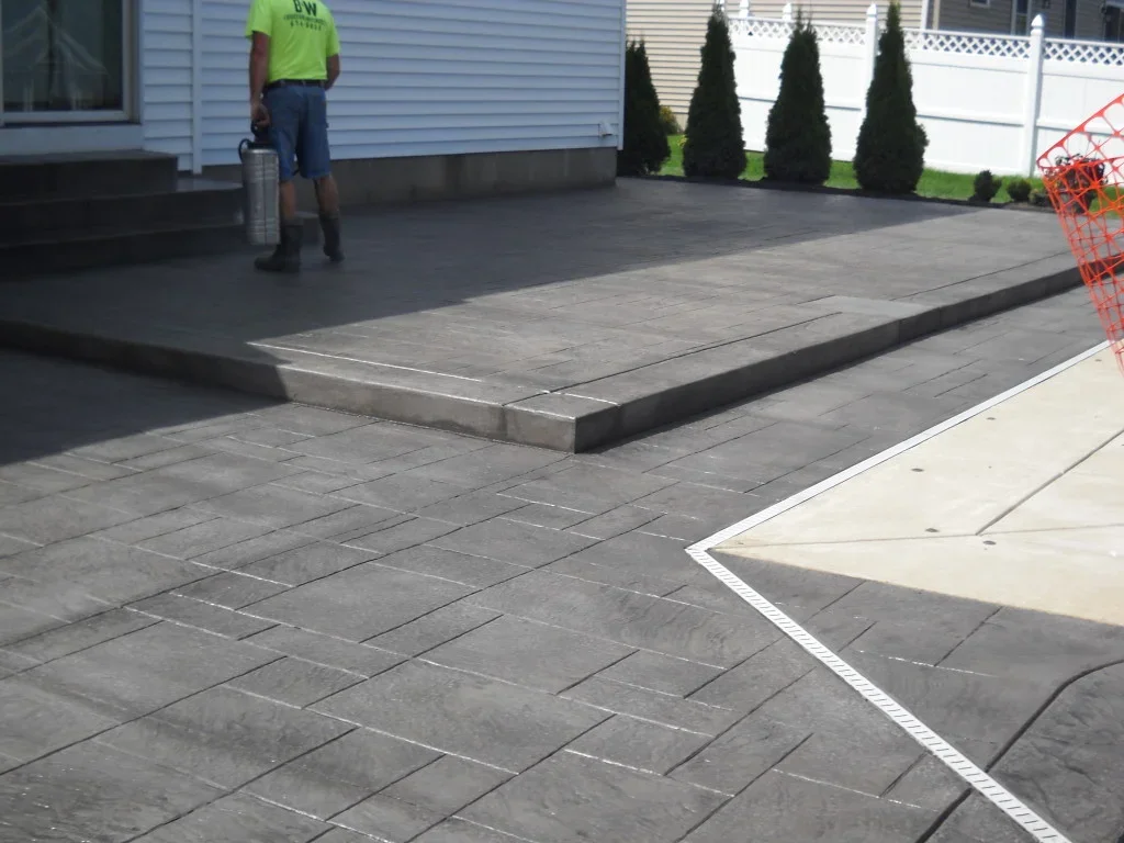 Paver installation on a backyard patio with a worker standing near the house holding a paver sealing or cleaning tool.