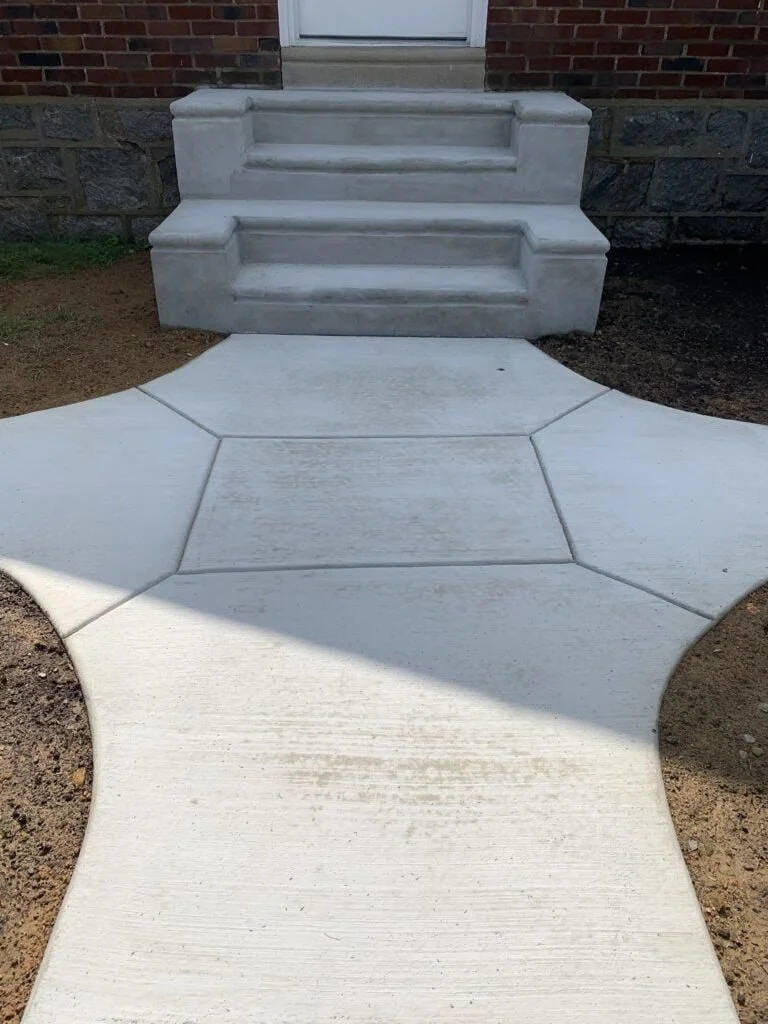 Concrete sidewalk leading to front steps of a house with a brick and stone exterior. The steps are made of concrete, and the sidewalk has a curved, decorative design.