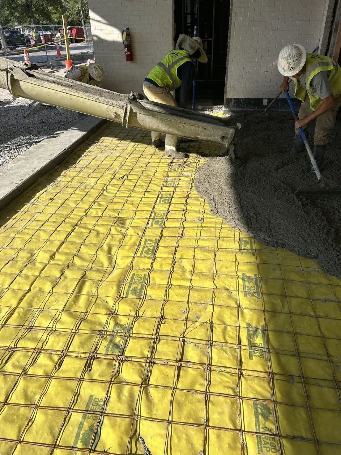 Construction workers pouring concrete on a sidewalk with yellow plastic reinforcement mesh.
