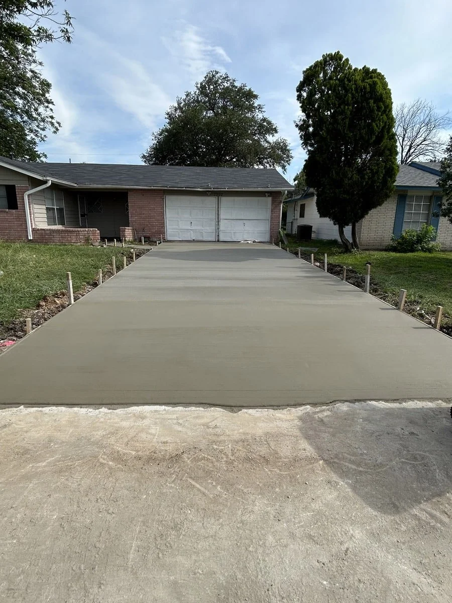 Newly poured concrete driveway leading to a garage in a residential neighborhood, with side posts marking the edges of the driveway, green grass on sides, and houses in the background.