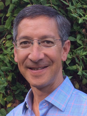 A man Roland Hsu with, glasses, and wearing a light blue checkered shirt, smiling in front of green foliage.