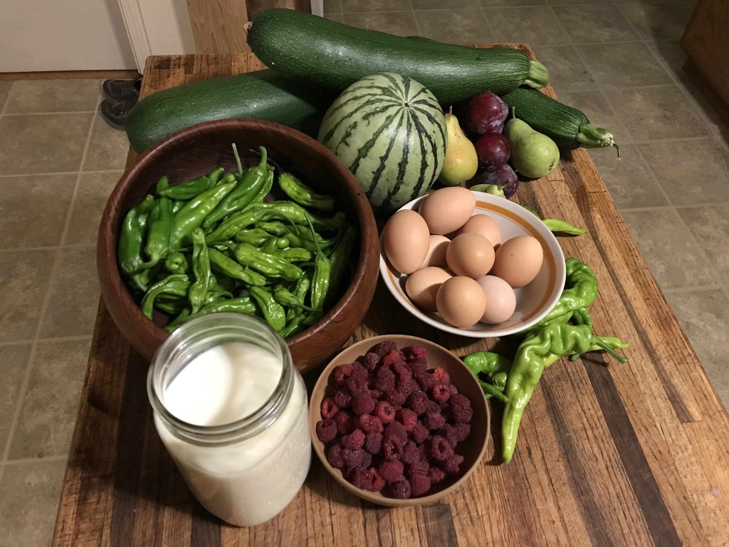 A rustic wooden table displays fresh vegetables, fruits, eggs, milk, and berries, including zucchinis, a watermelon, red and green apples, grapes, a bowl of raspberries, a bowl of eggs, and a jar of milk.
