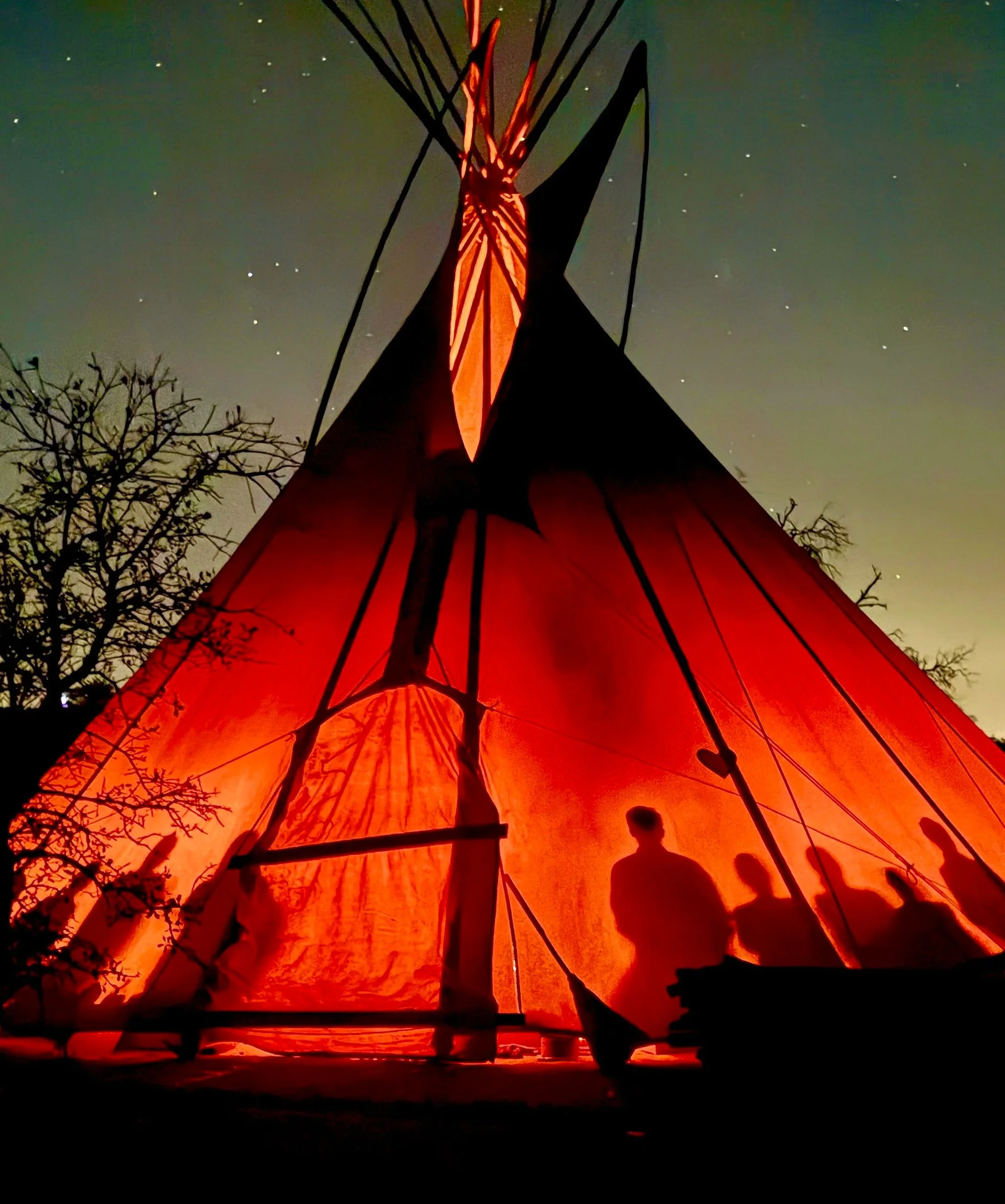 Silhouette of two people standing near a small boat under a starry night sky, with an illuminated orange fabric or structure in the background.