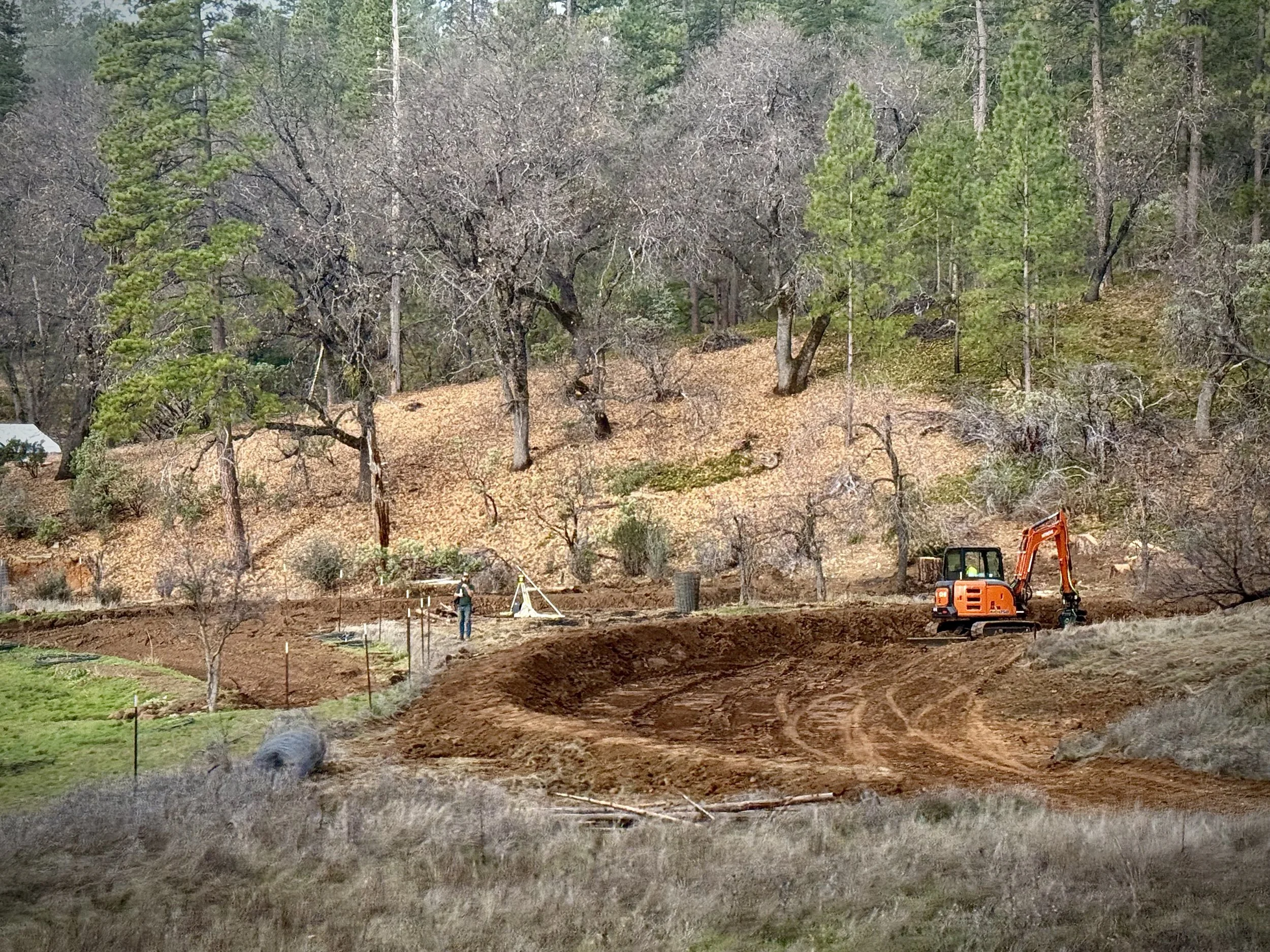 Construction site with a small orange excavator and workers, surrounded by trees, dirt, and landscaping equipment.