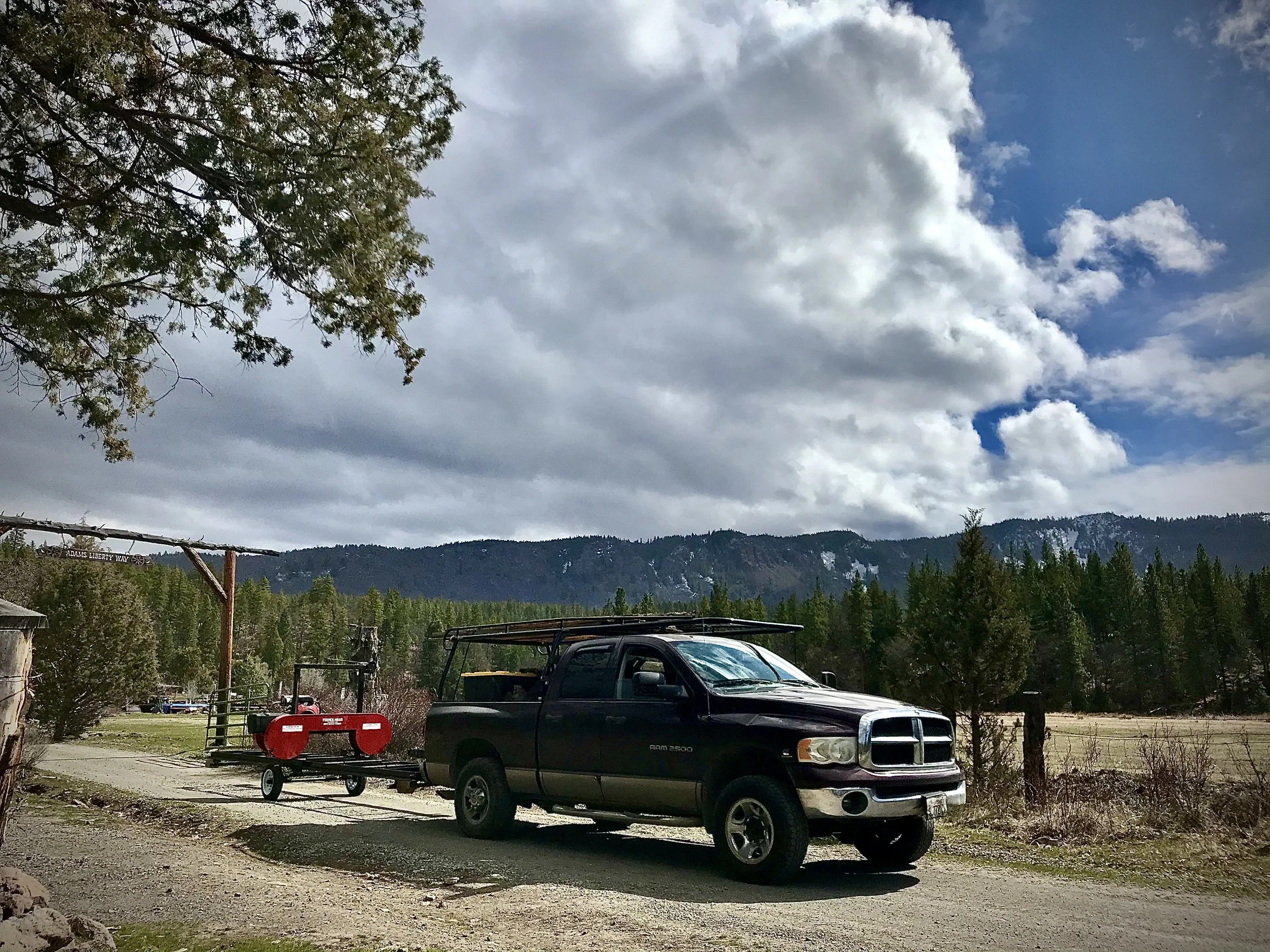 A rural scene showing a black pickup truck parked on a dirt driveway with a small red trailer hitched to it, a wooden sign that says 'ADAMS LIBERTY WAY,' and a backdrop of mountains with partly cloudy skies and some trees.